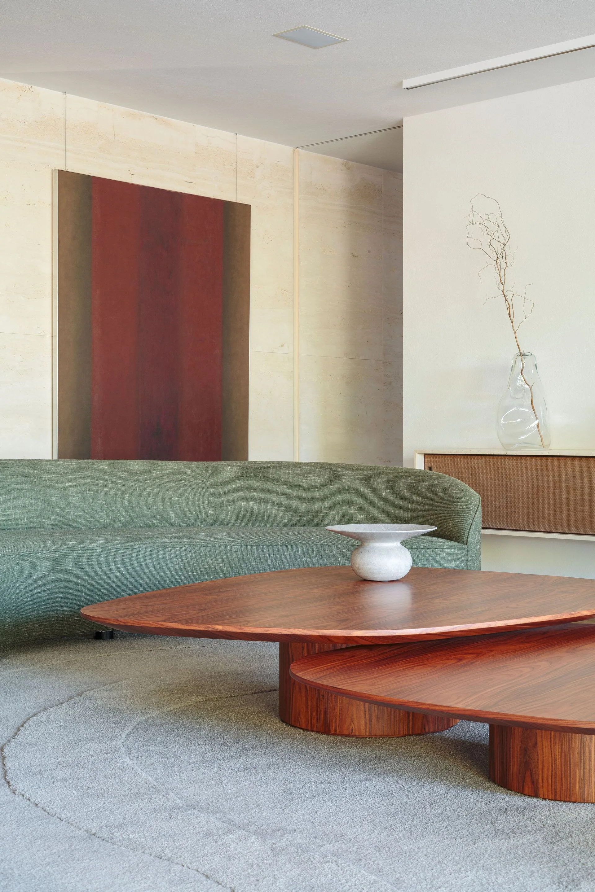 A living room featuring a curved green sofa, organic wood nesting coffee tables, a textured stone wall, and a large abstract painting.