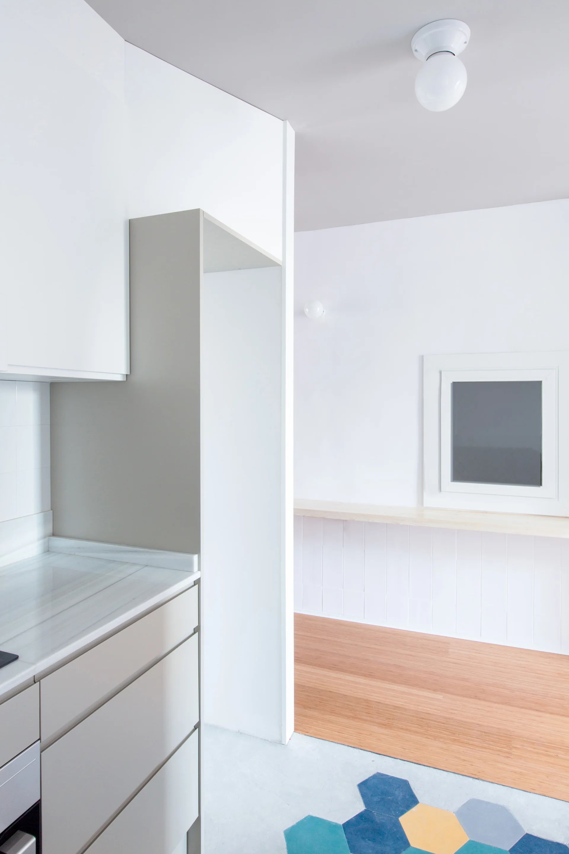 Minimalist kitchen featuring matte taupe cabinets, white countertops, and colorful hexagonal floor tiles transitioning into wood flooring.