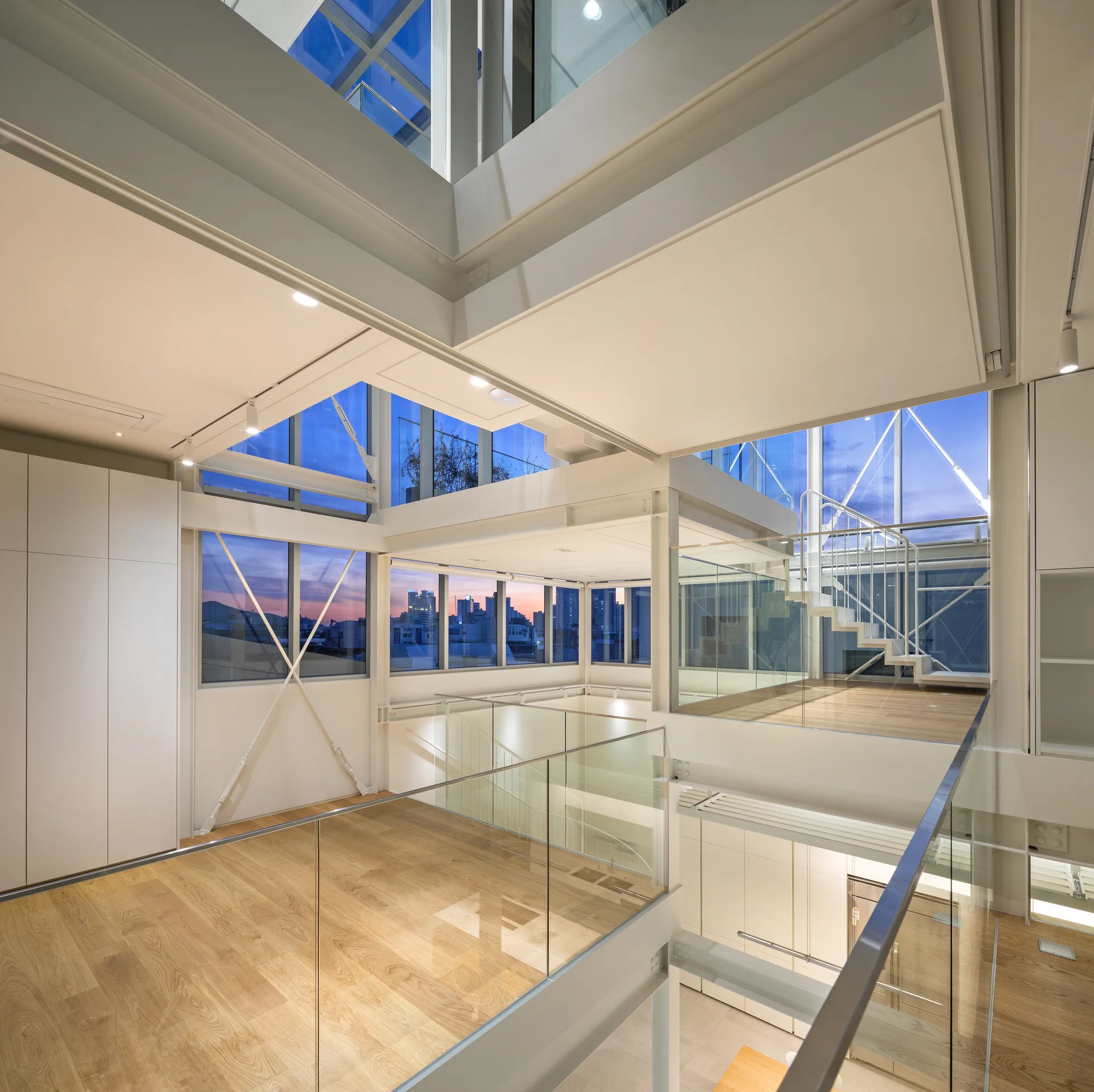 A modern multi-level interior featuring open mezzanine levels with glass railings, light wood flooring, and structural white steel beams under a skylight.