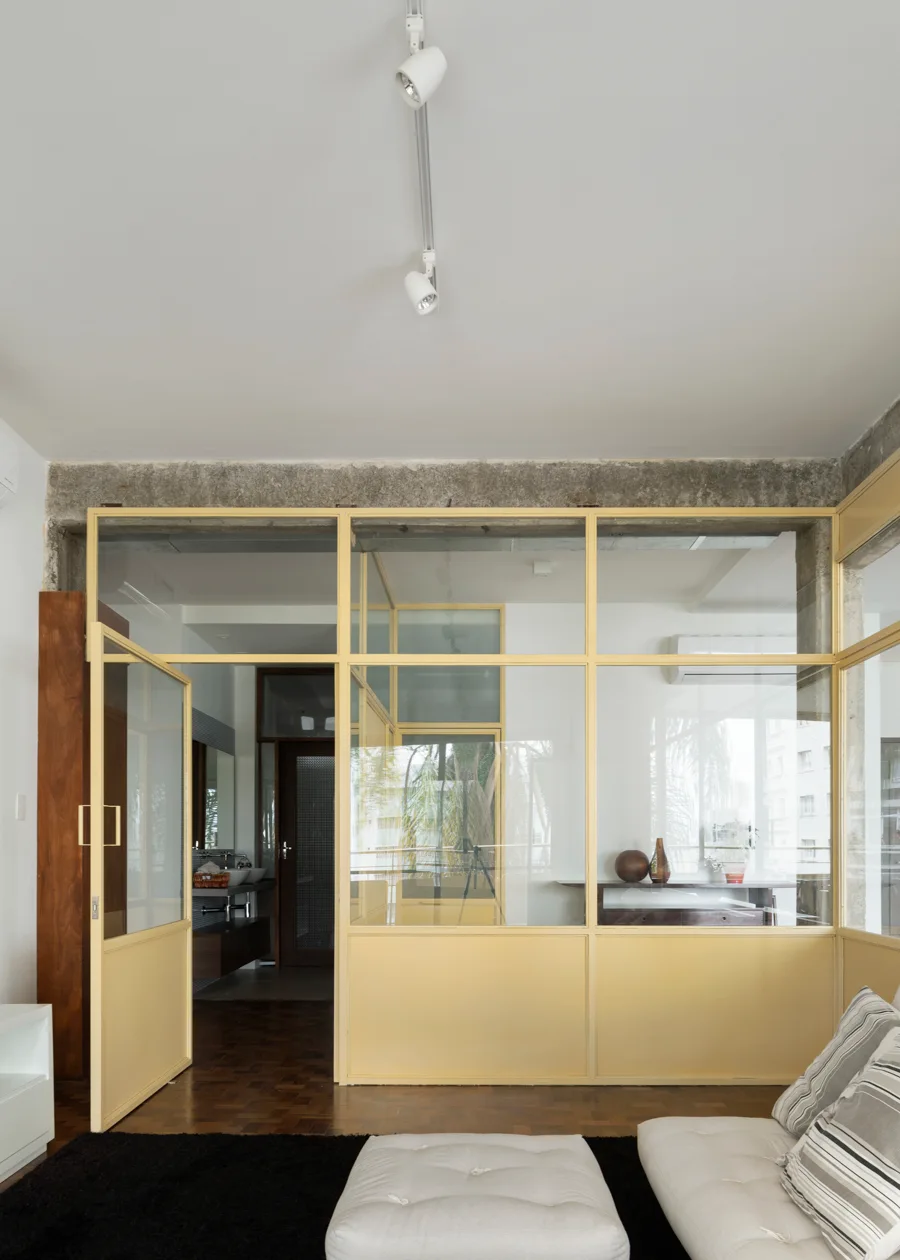 A modern living space features a yellow-framed glass partition wall, an exposed concrete ceiling beam, dark parquet floors, and a white sofa.