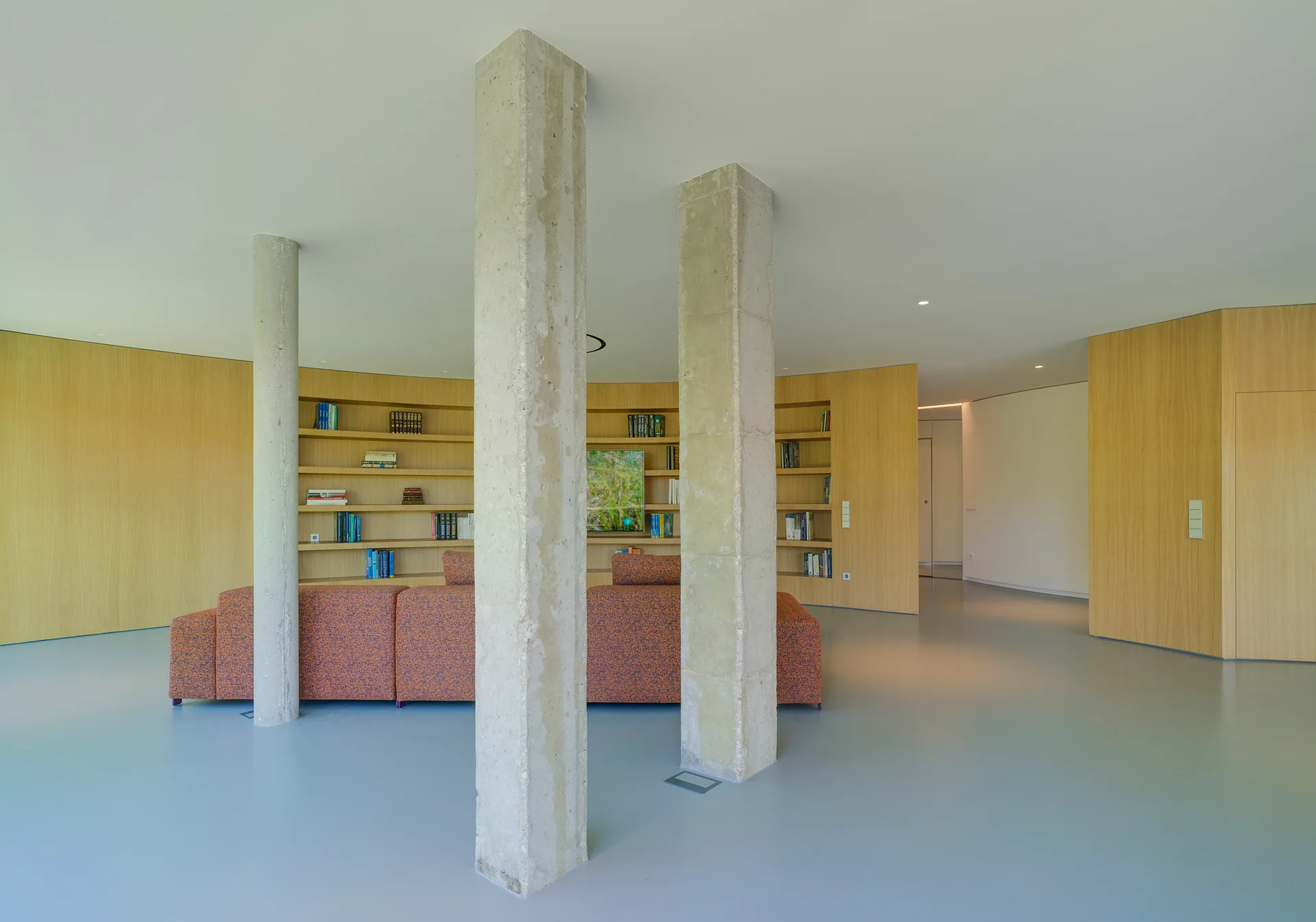 A minimalist open-plan living room featuring exposed raw concrete columns, a curved wood paneled bookshelf wall, and a textured orange sofa on a grey floor.