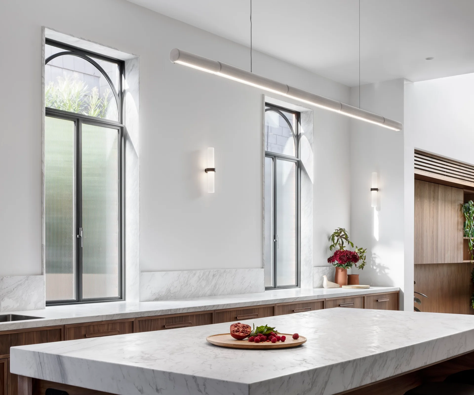 A modern kitchen features a large white marble island, wood base cabinets, two arched windows with textured reeded glass, and a long linear pendant light.