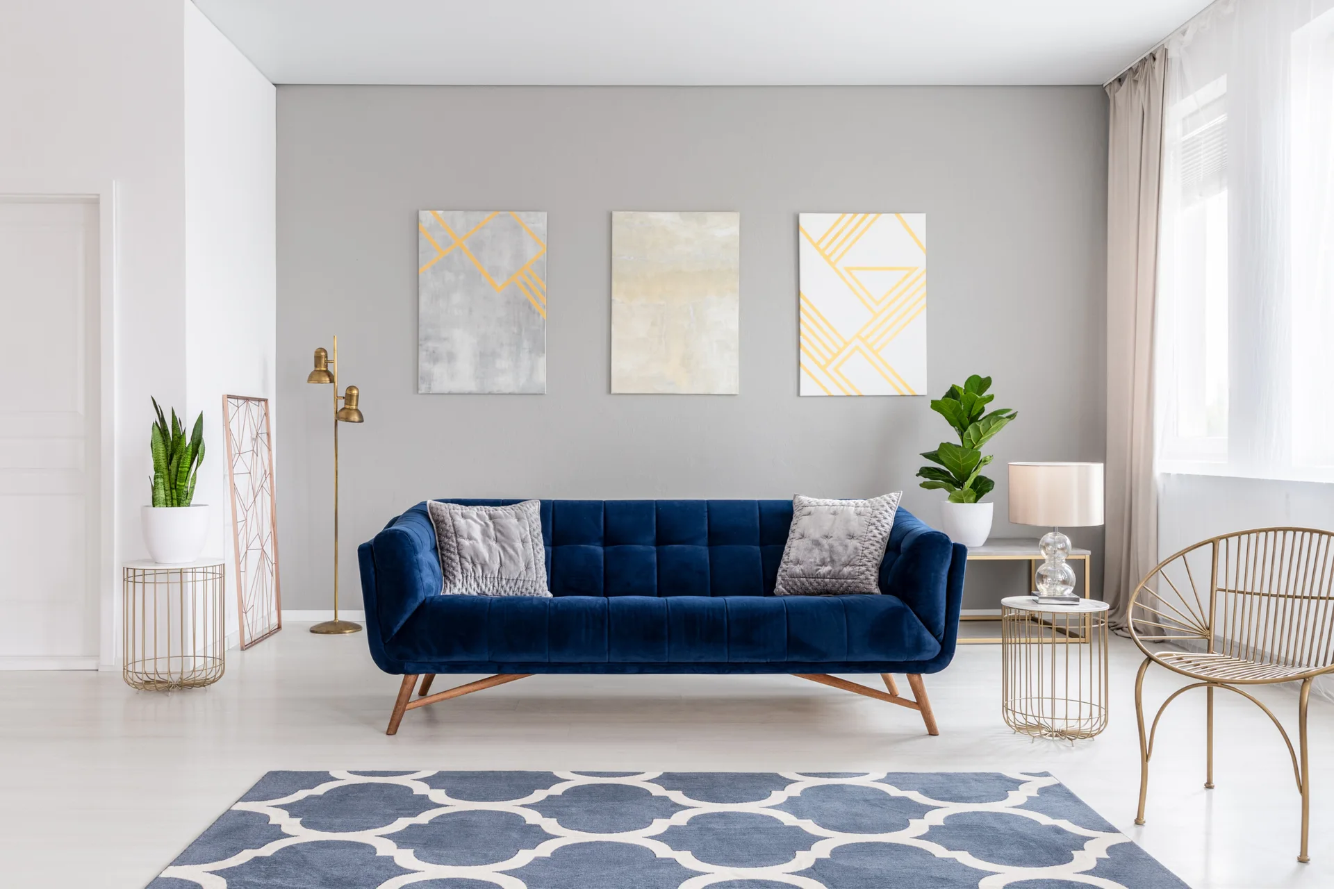 A contemporary living room features a blue velvet sofa, a gray accent wall with abstract paintings, gold wire side tables, and a patterned blue rug.