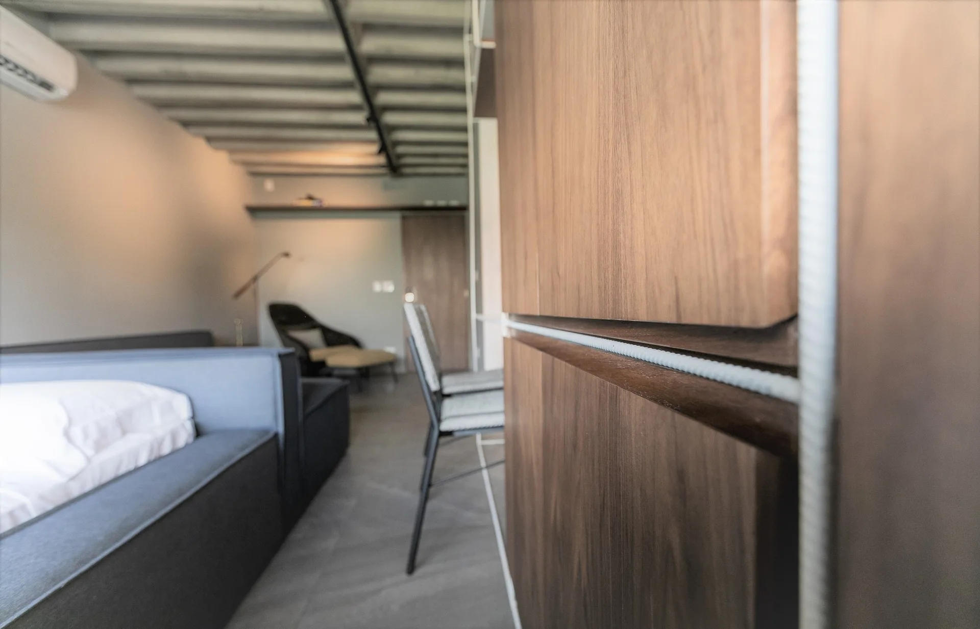 A modern studio apartment features a prominent wood-toned cabinet in the foreground, a grey sofa, and an exposed concrete beam ceiling.