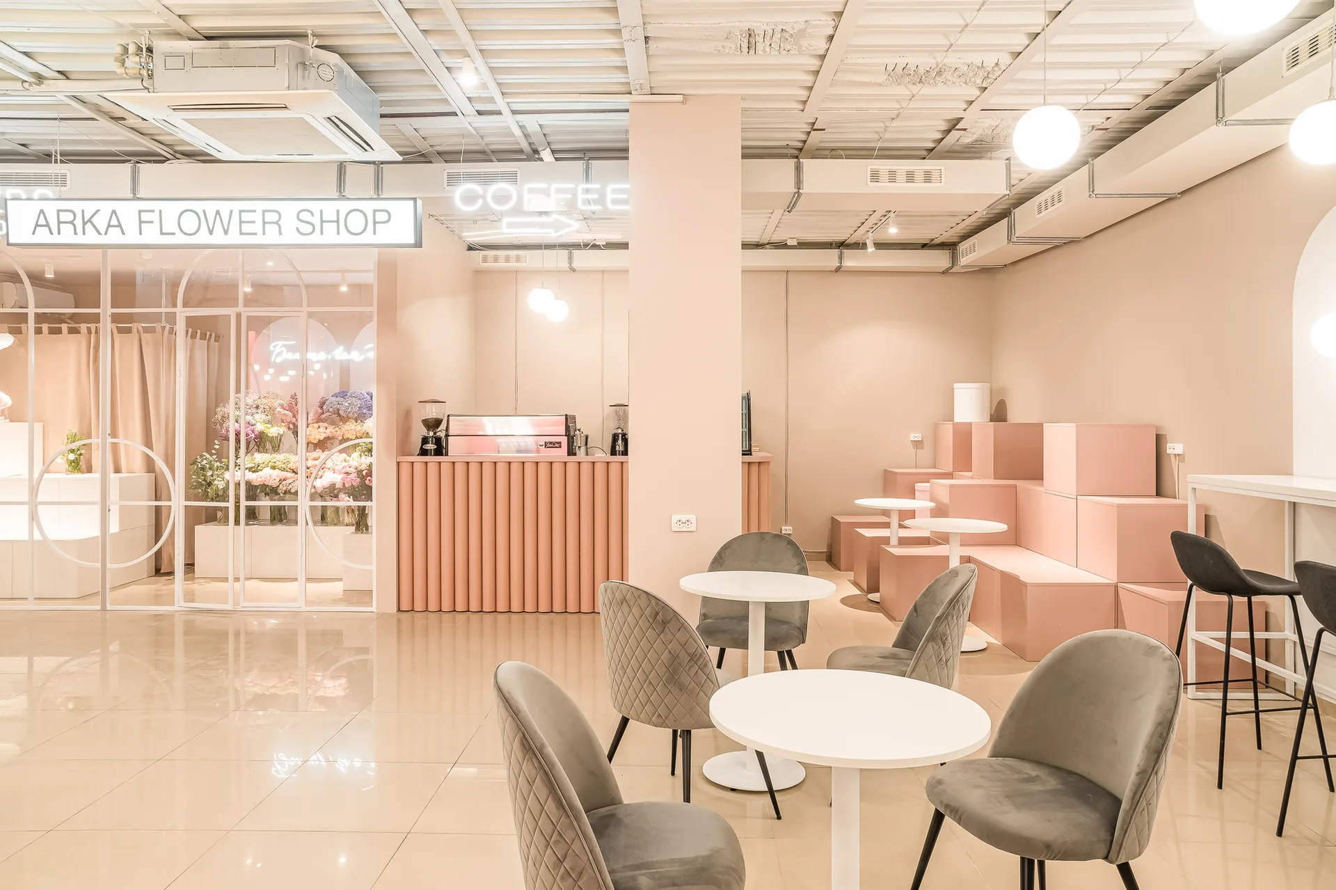 A bright pink cafe interior featuring grey upholstered chairs, white bistro tables, a fluted service counter, and a glass-enclosed flower shop.