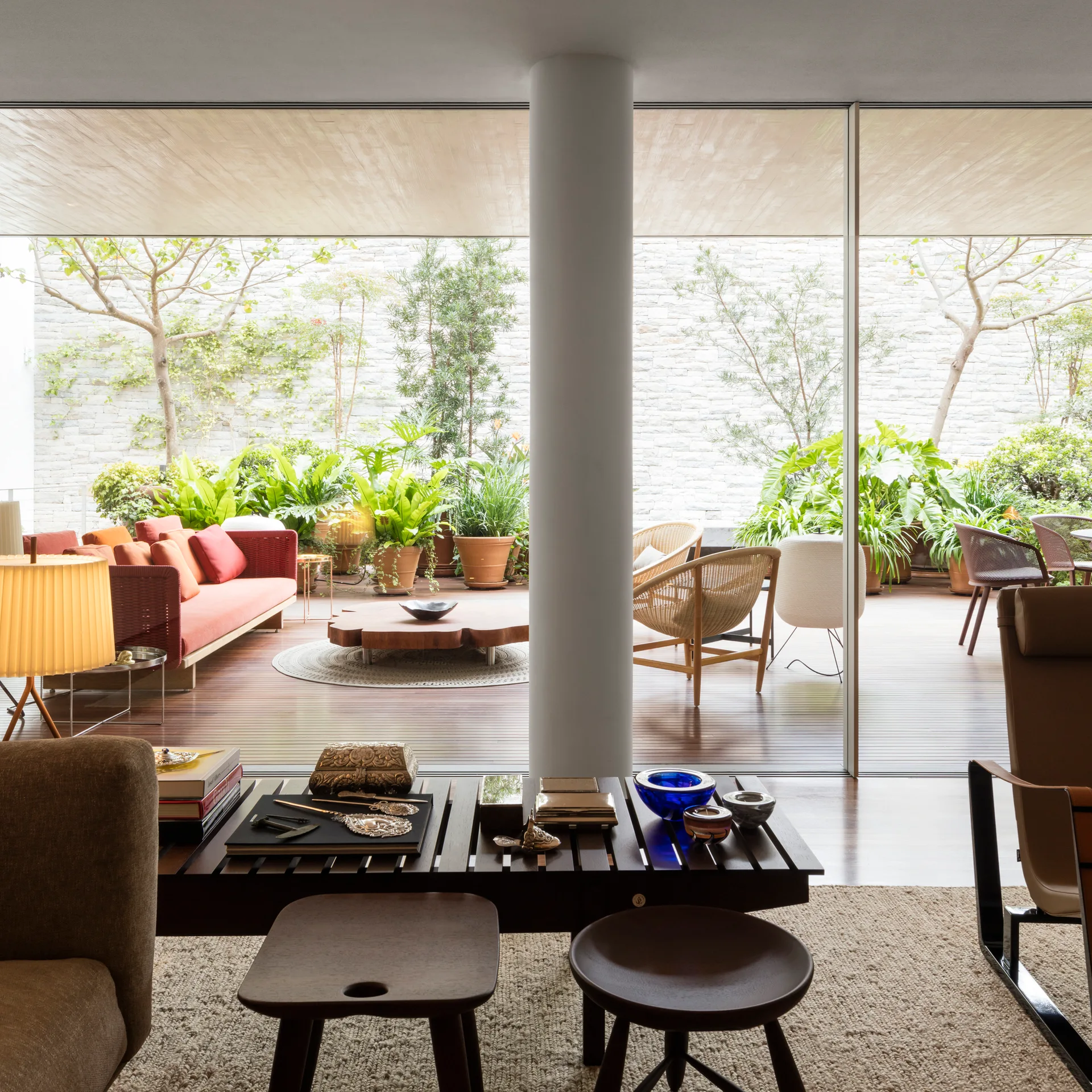 An indoor living room with wood stools in the foreground looks out to a plant-filled deck with a terracotta sofa through large sliding glass doors.