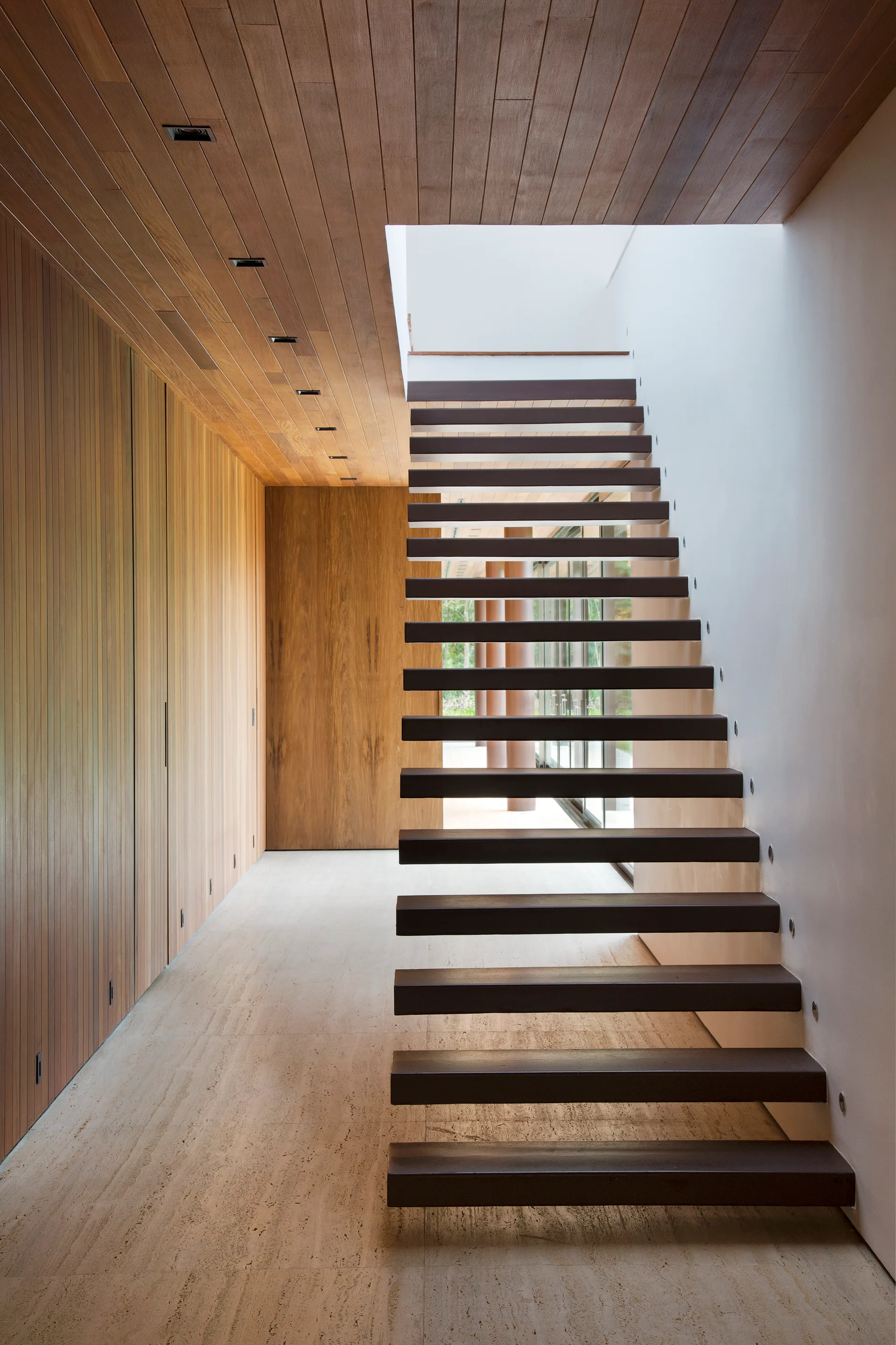 A modern hallway features a floating wooden staircase next to a vertical wood slat wall, matching wood ceiling, and cream stone flooring.