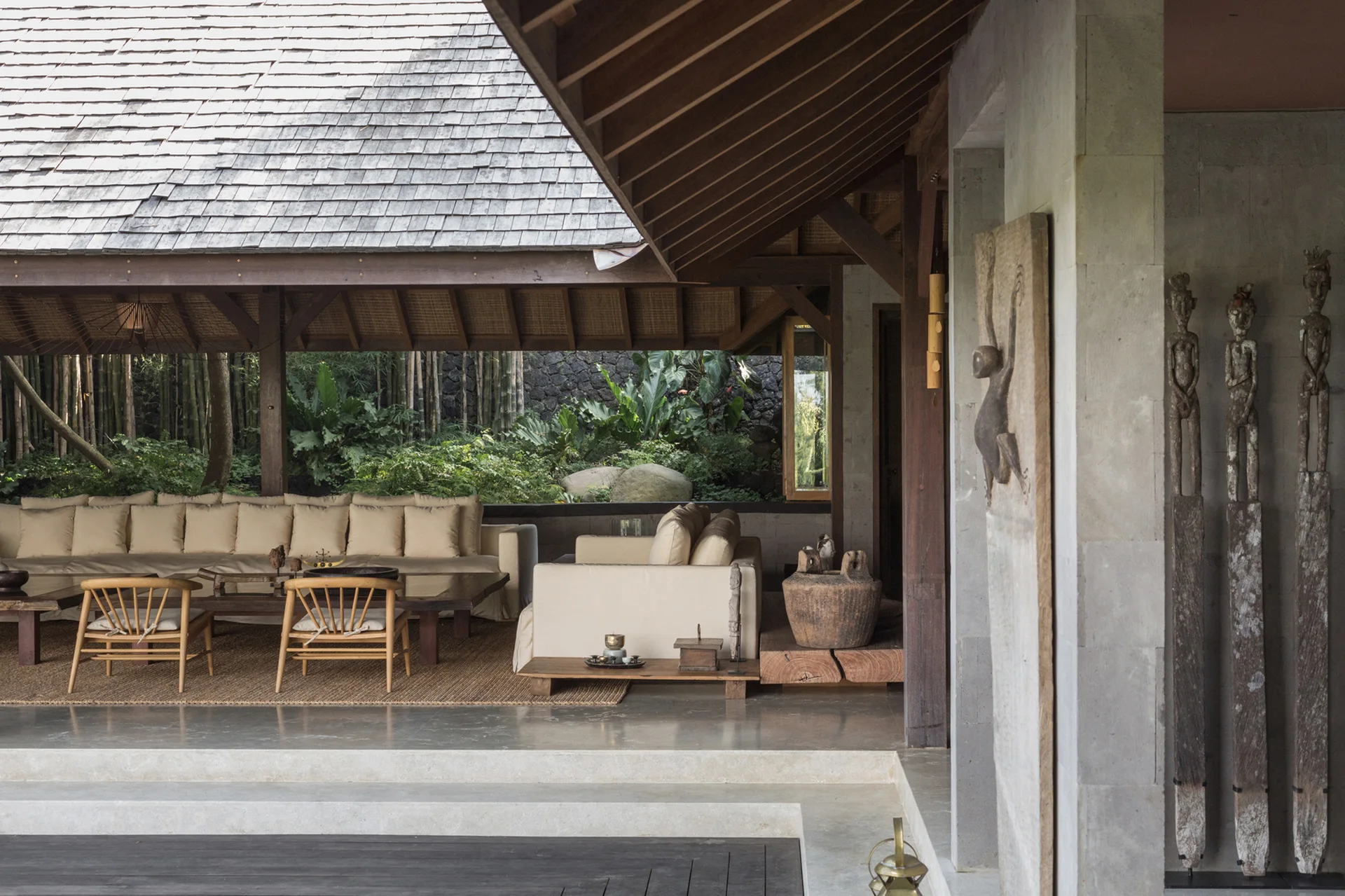 An open-air pavilion lounge features low light-colored sofas, wooden spindle chairs, and tall carved statues against a lush green background.