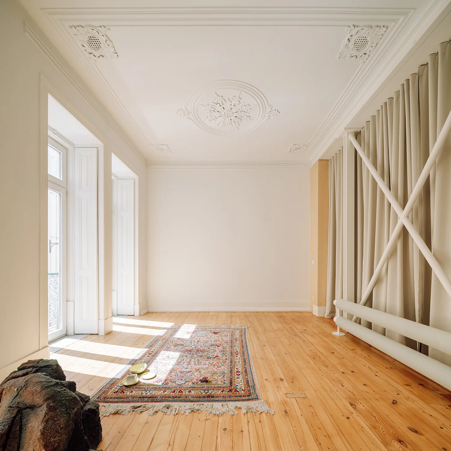 An empty room features detailed plaster ceiling medallions, light wood flooring, a patterned runner rug, and tall windows framed by beige drapery.