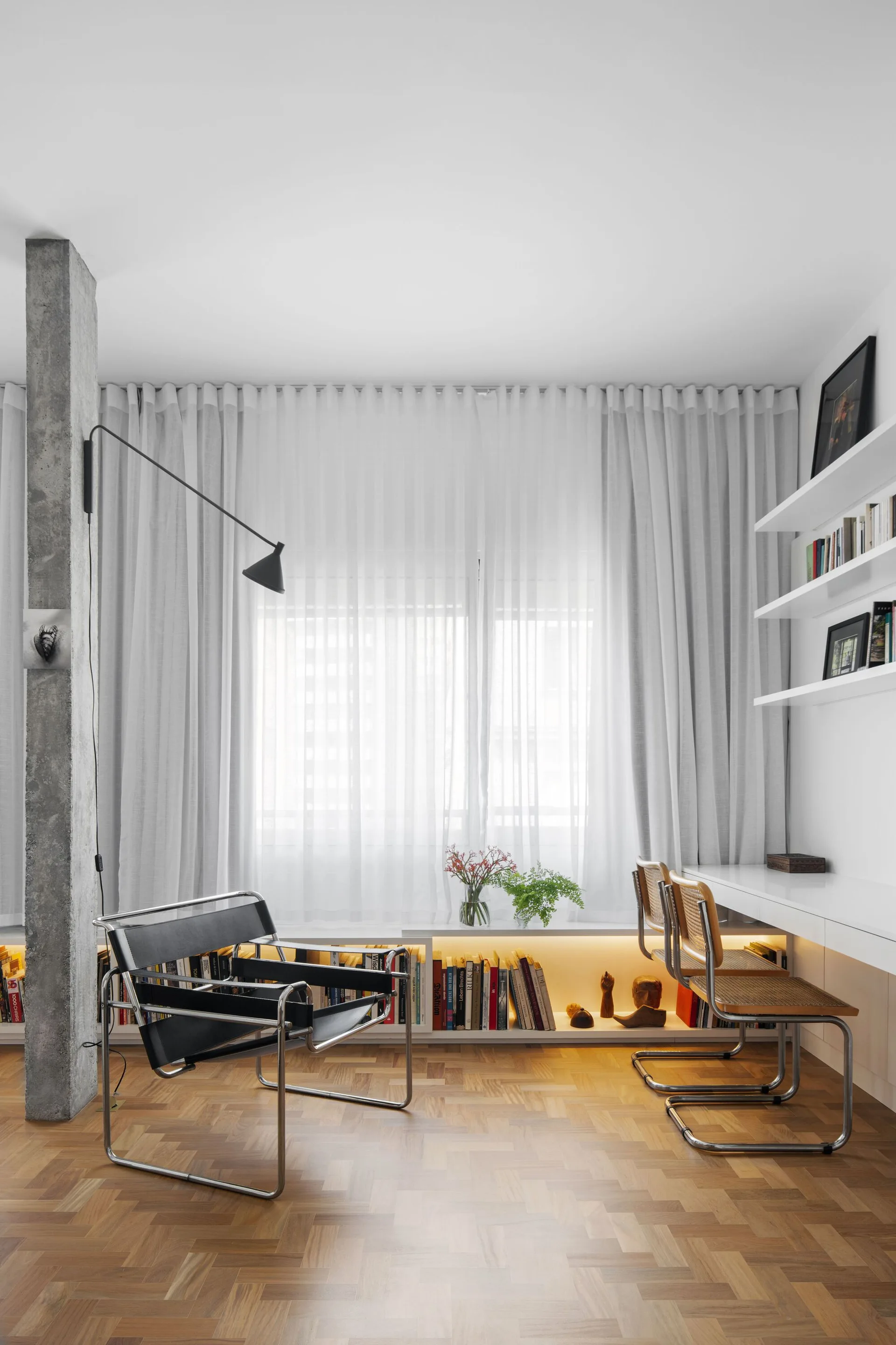 A modern study displays a white built-in desk with cane chairs, a black leather armchair, and a low bookshelf illuminated by warm integrated lighting.
