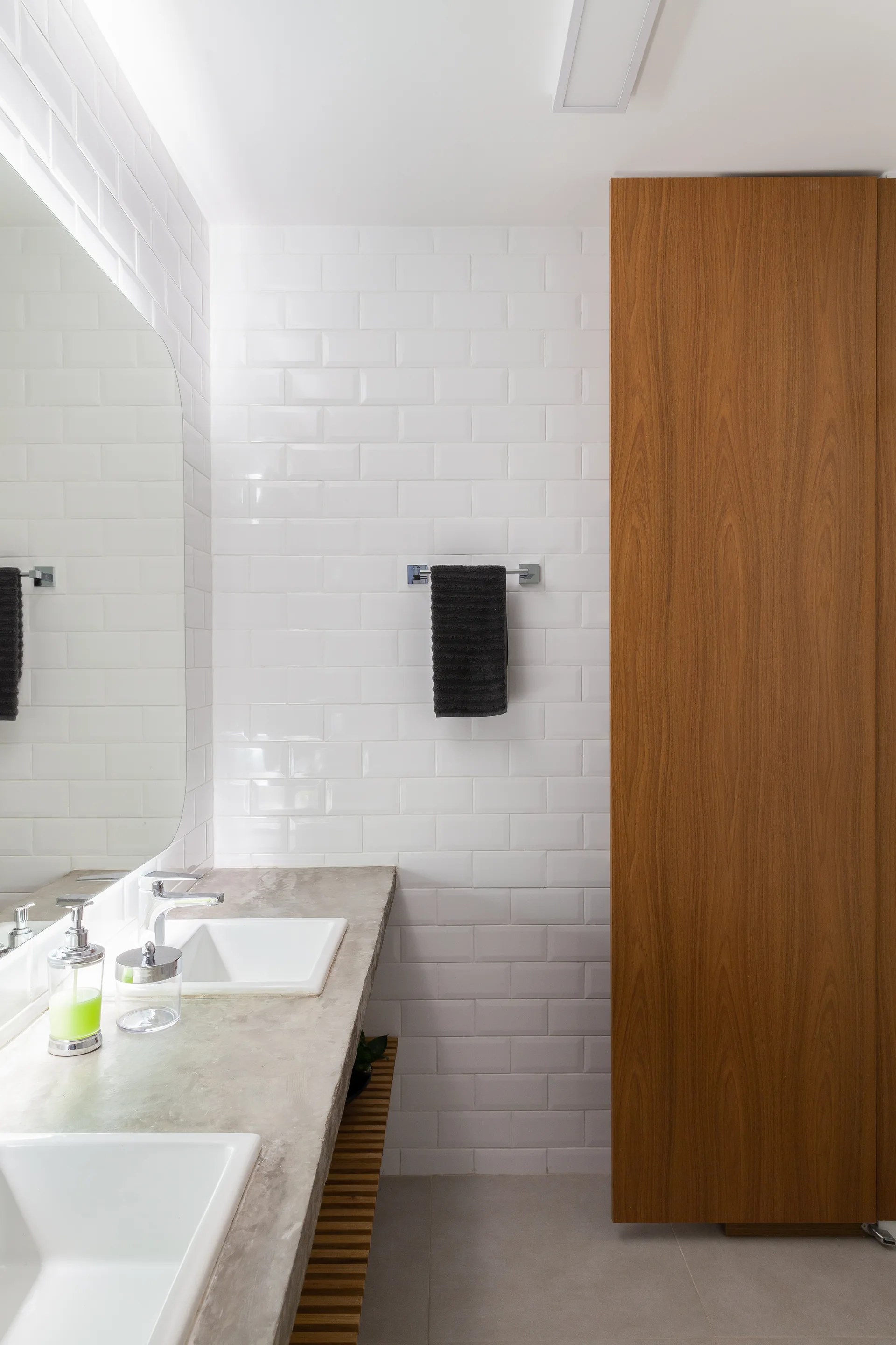 A modern bathroom vanity with a concrete countertop, double sinks, a glossy white subway tile wall, and a vertical wood door.