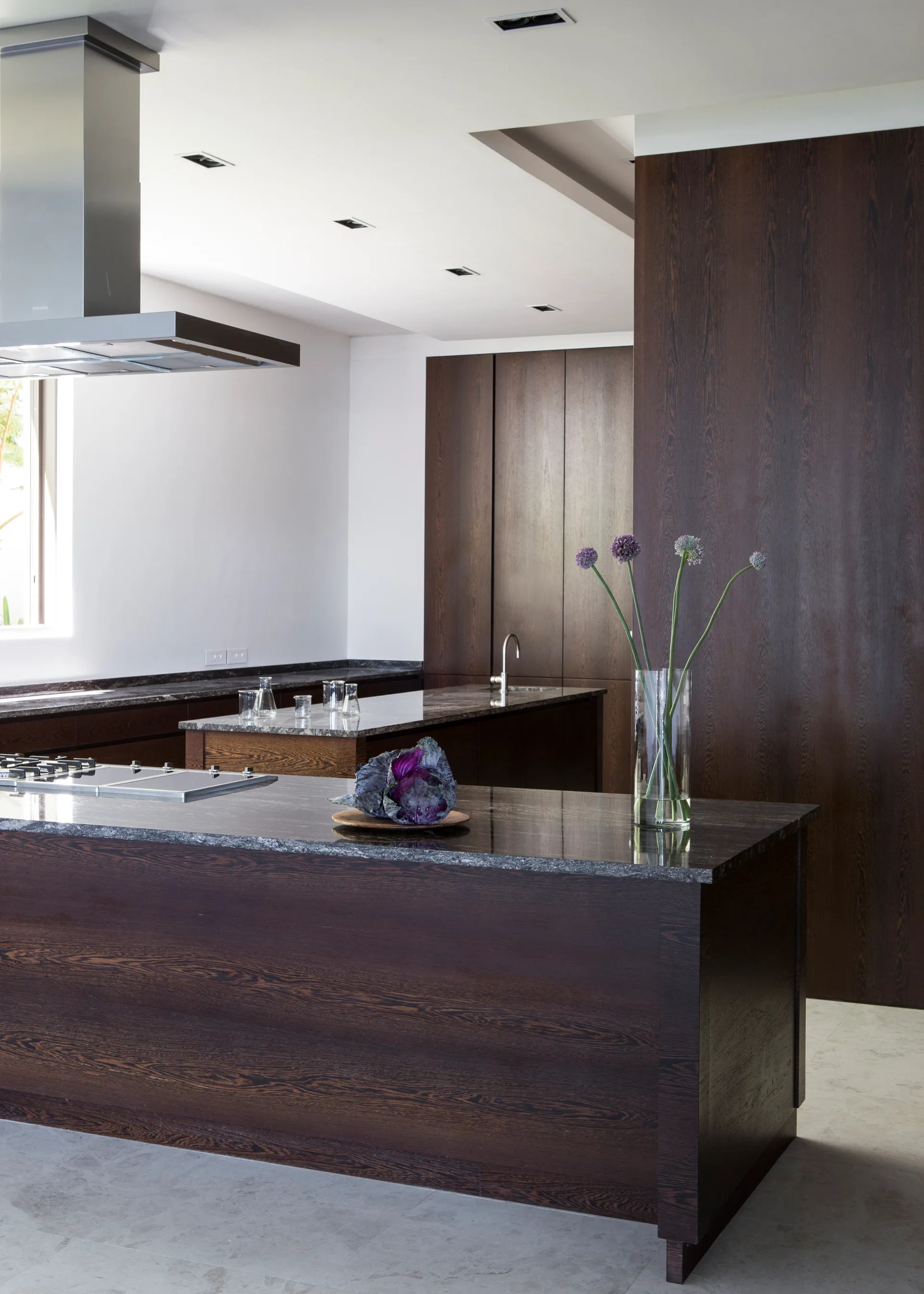 A modern kitchen features dark brown wood cabinetry, double islands with dark stone countertops, and a stainless steel range hood.
