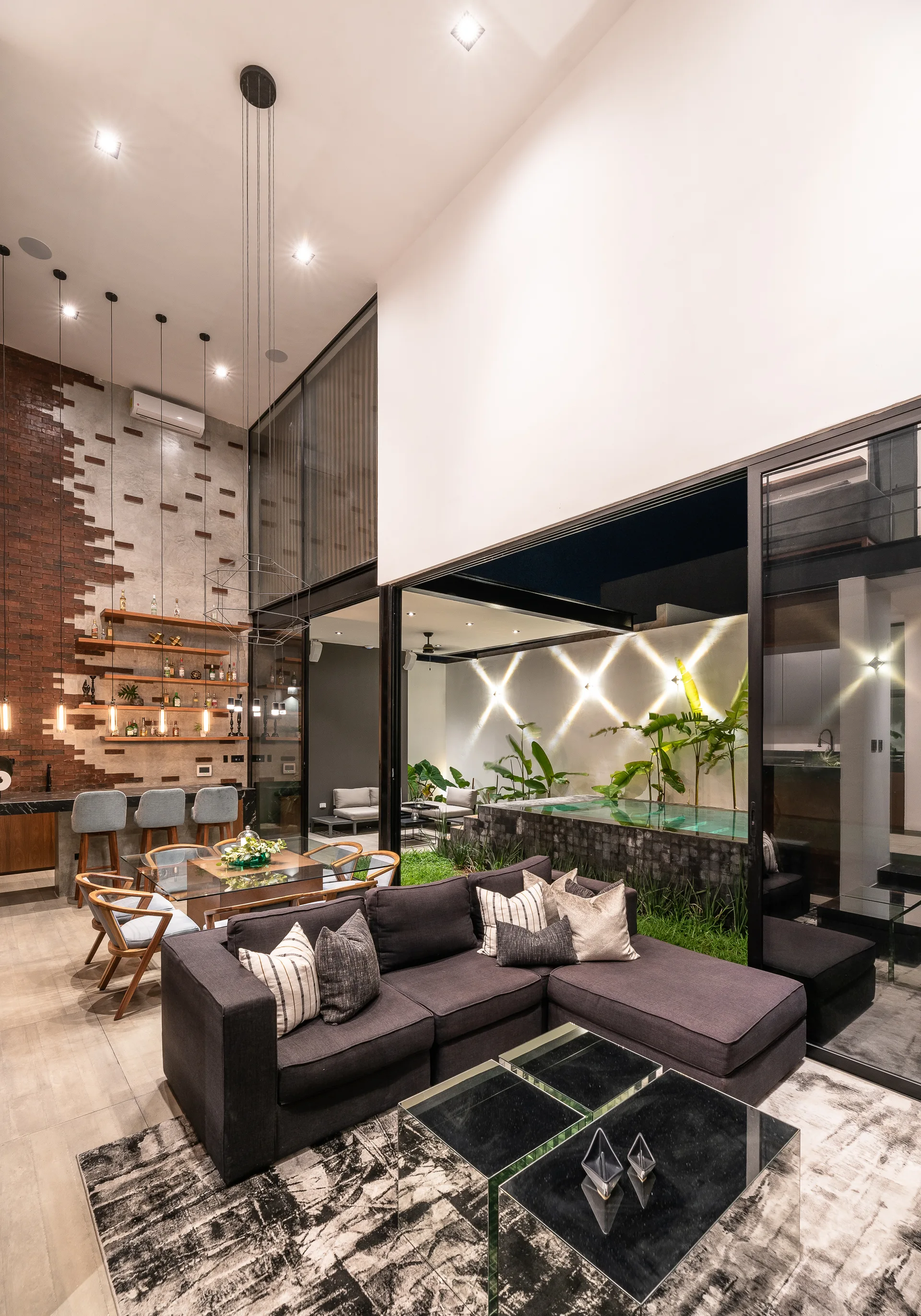 A grey sectional sofa sits on a textured rug next to a dining area, with glass walls opening to a courtyard pool and garden.