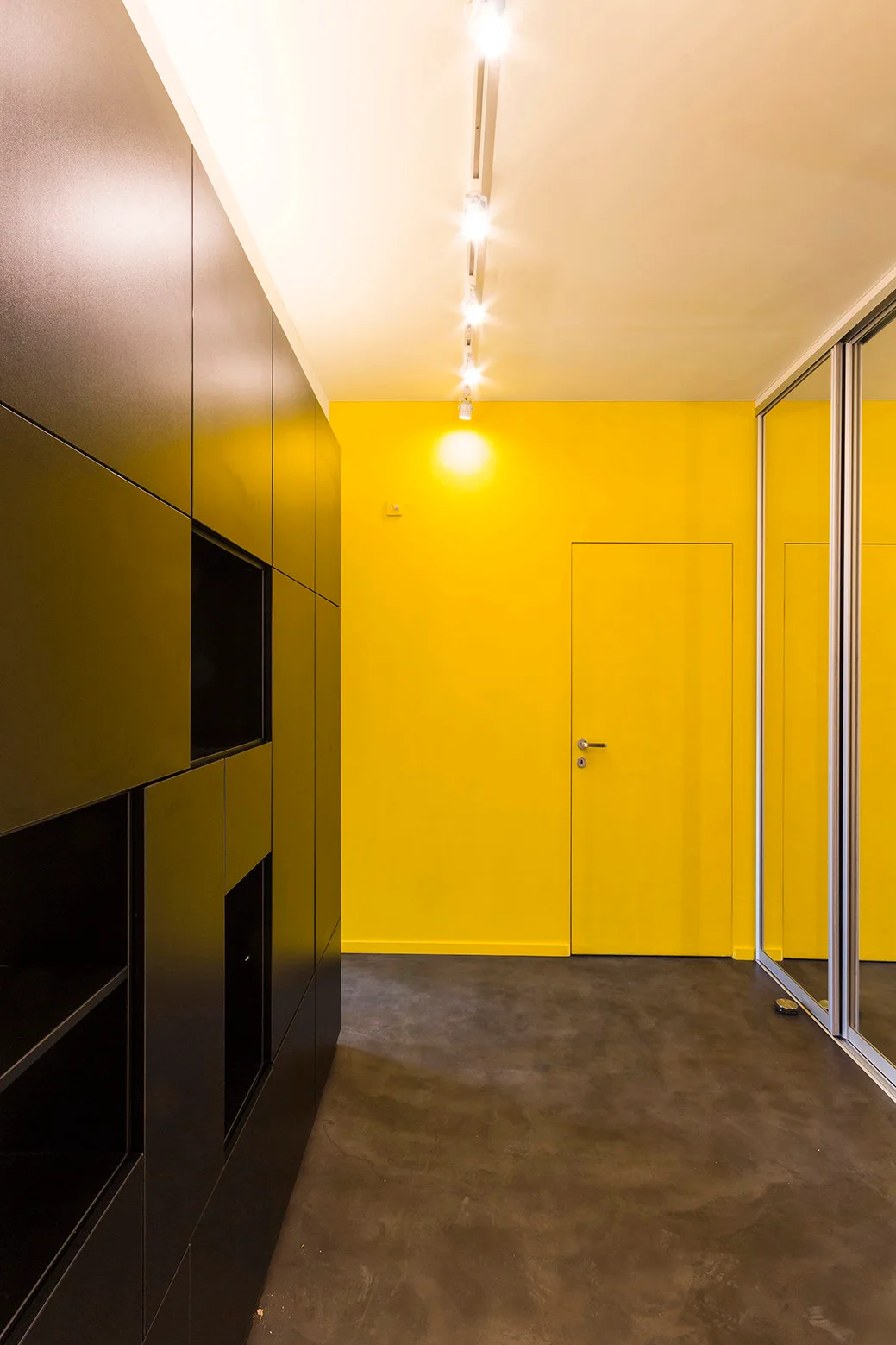 A modern hallway features a bright yellow wall with an integrated door, matte black storage cabinets, and mirrored closet doors.