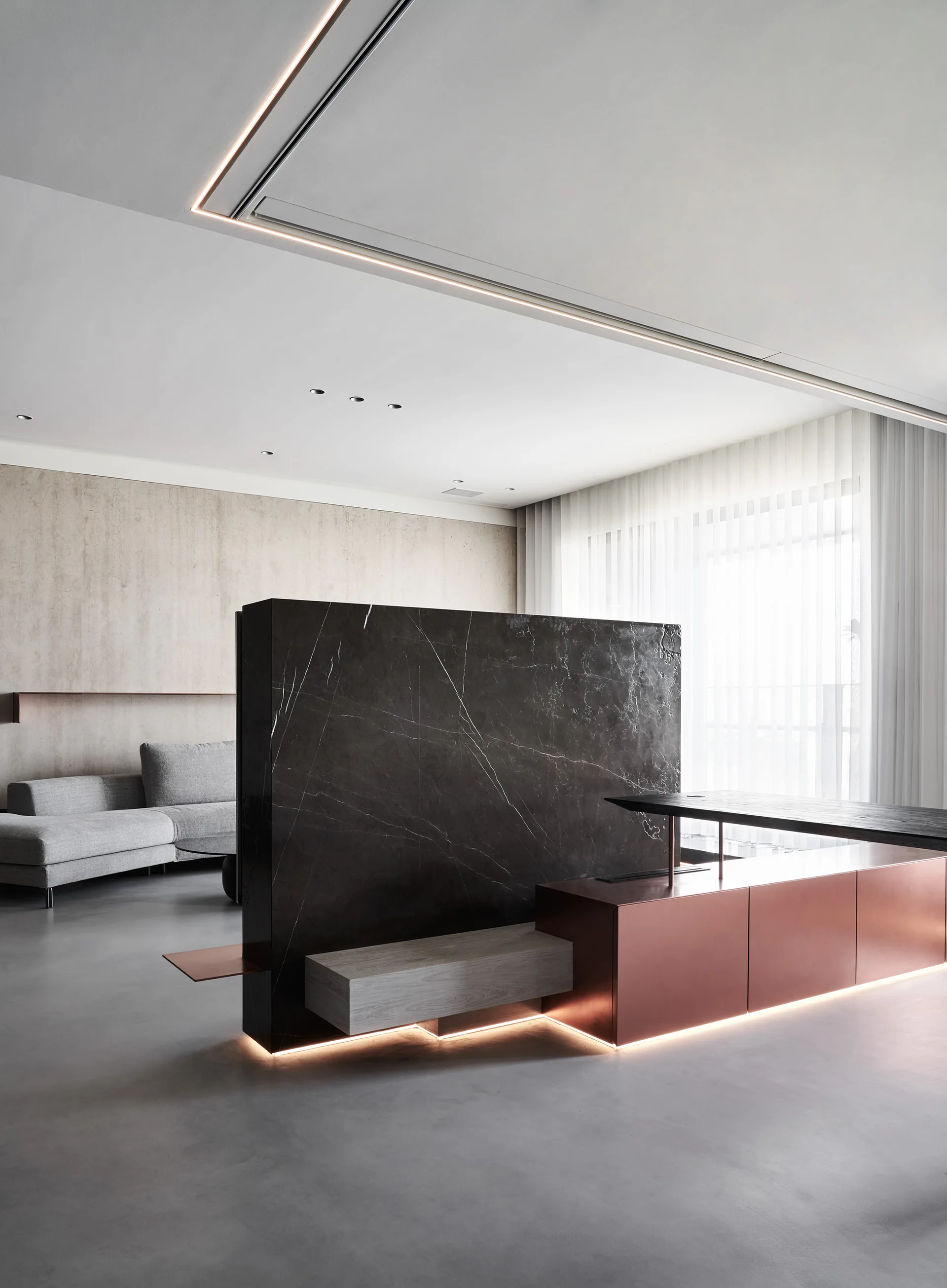 A dark marble partition wall with copper base cabinets and recessed floor lighting stands in a minimalist living room with a grey sofa and sheer curtains.