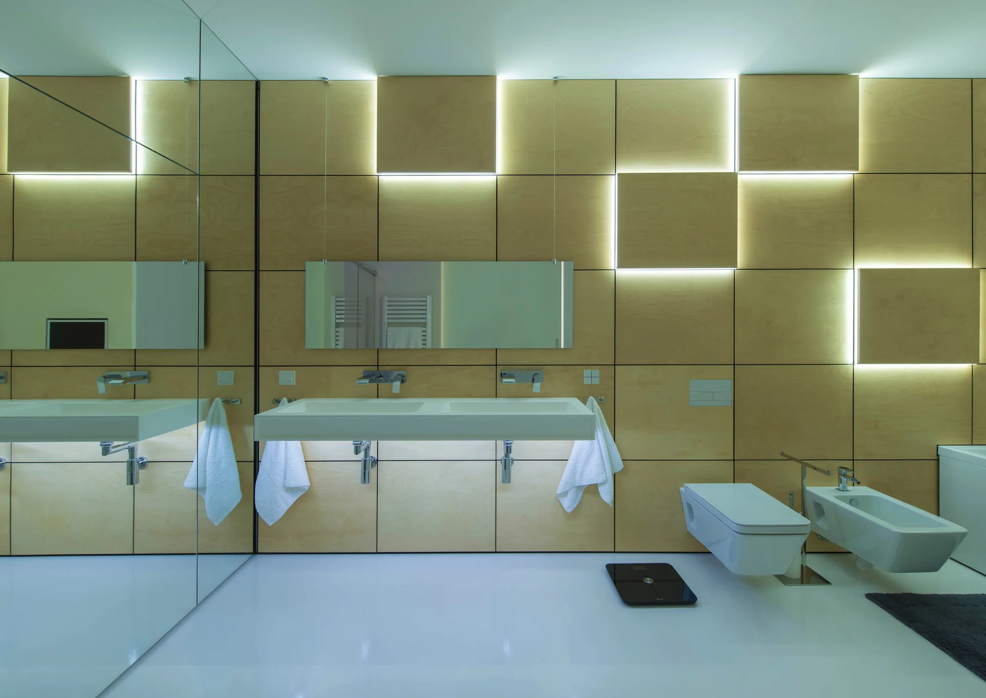 A minimalist bathroom features a floating white double sink, wall-mounted fixtures, and light wood wall panels with integrated glowing square LED lights.