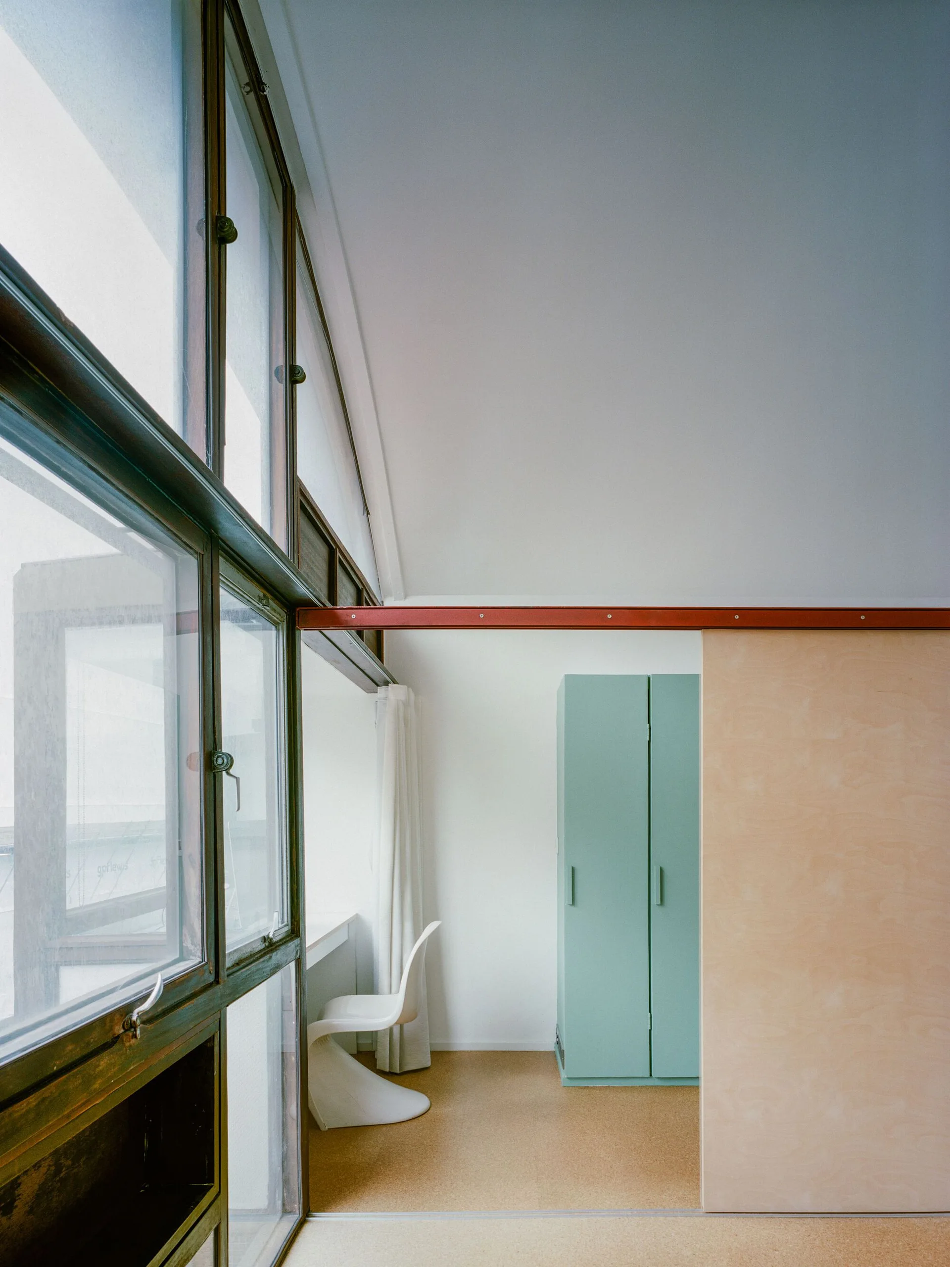 A minimalist modernist room featuring a prominent red structural beam, a light green cabinet, a plywood sliding door, and a white chair.