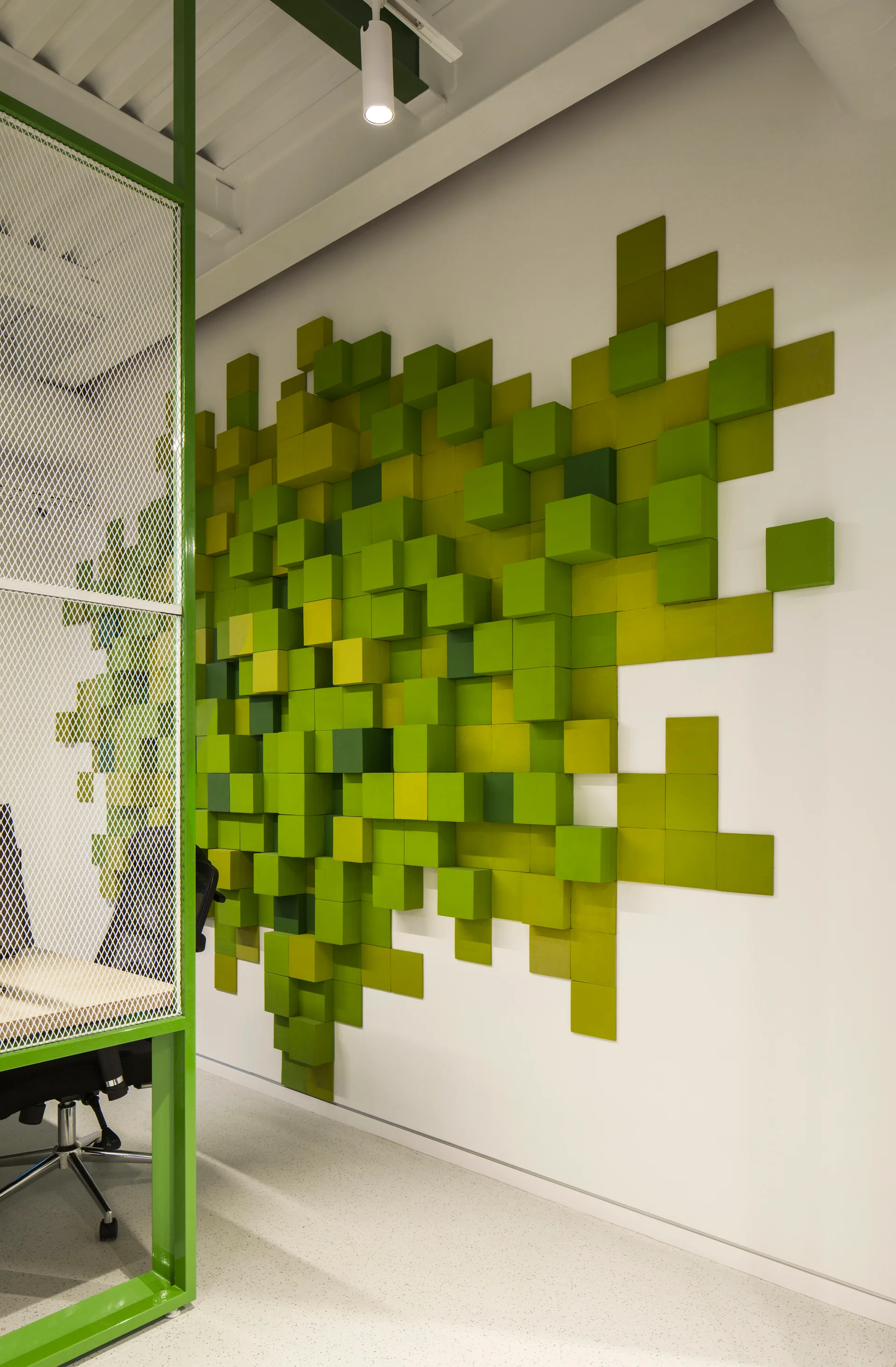 A white workspace featuring a three-dimensional green modular block wall installation next to a green metal and white mesh office partition.