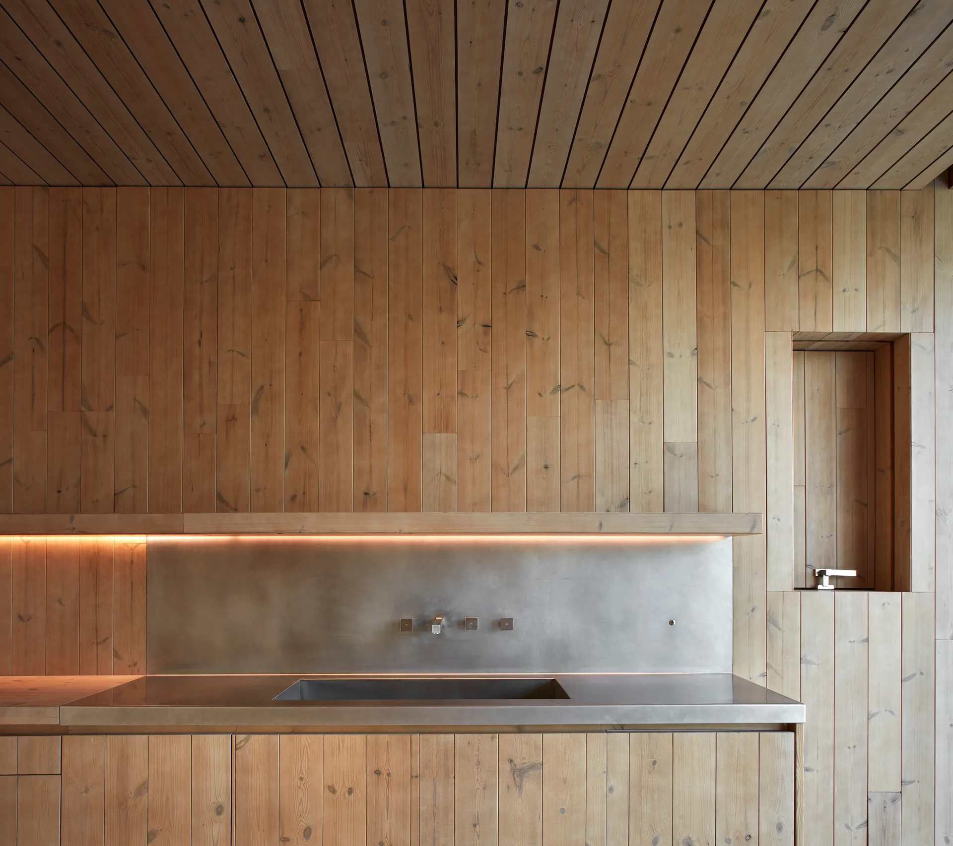 A minimalist kitchen features vertical wood paneling on the walls, a stainless steel countertop with an integrated sink, and an illuminated shelf.