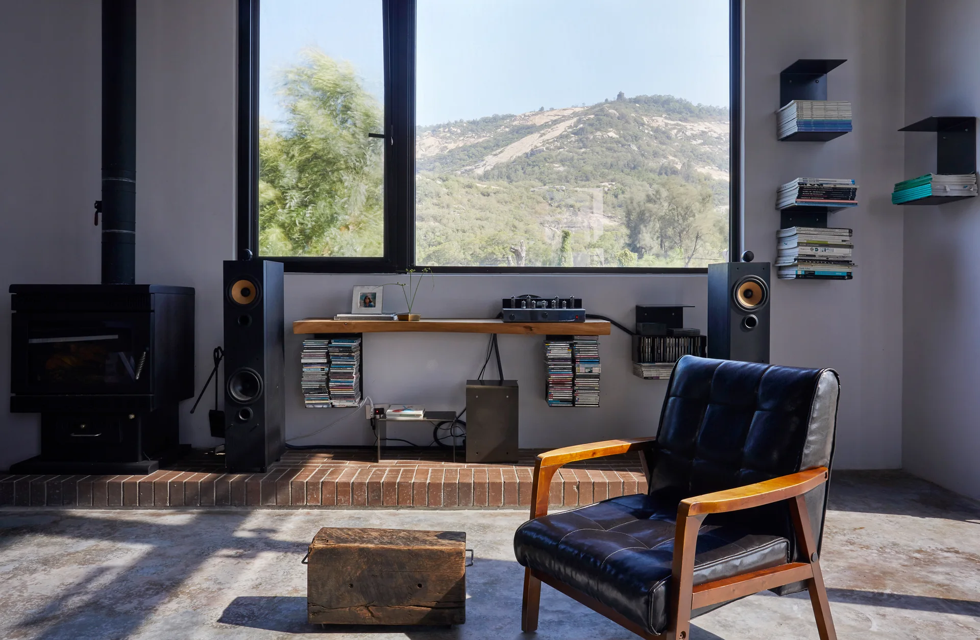 A minimalist living space featuring a black leather armchair, a floating live-edge wood console under a large window, and floorstanding speakers.