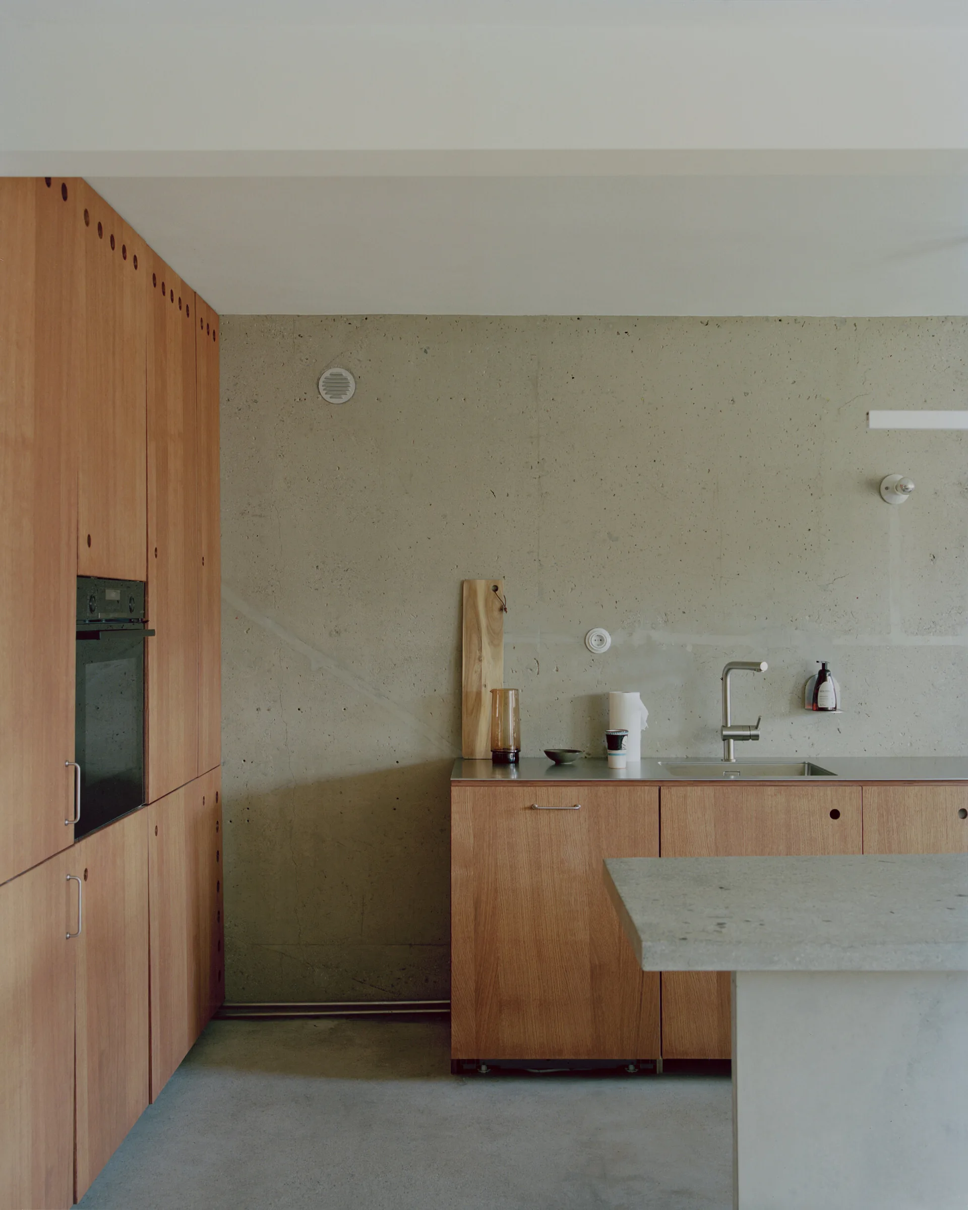 A minimalist kitchen features warm wood cabinetry, an exposed concrete wall, a stainless steel sink countertop, and a concrete island.
