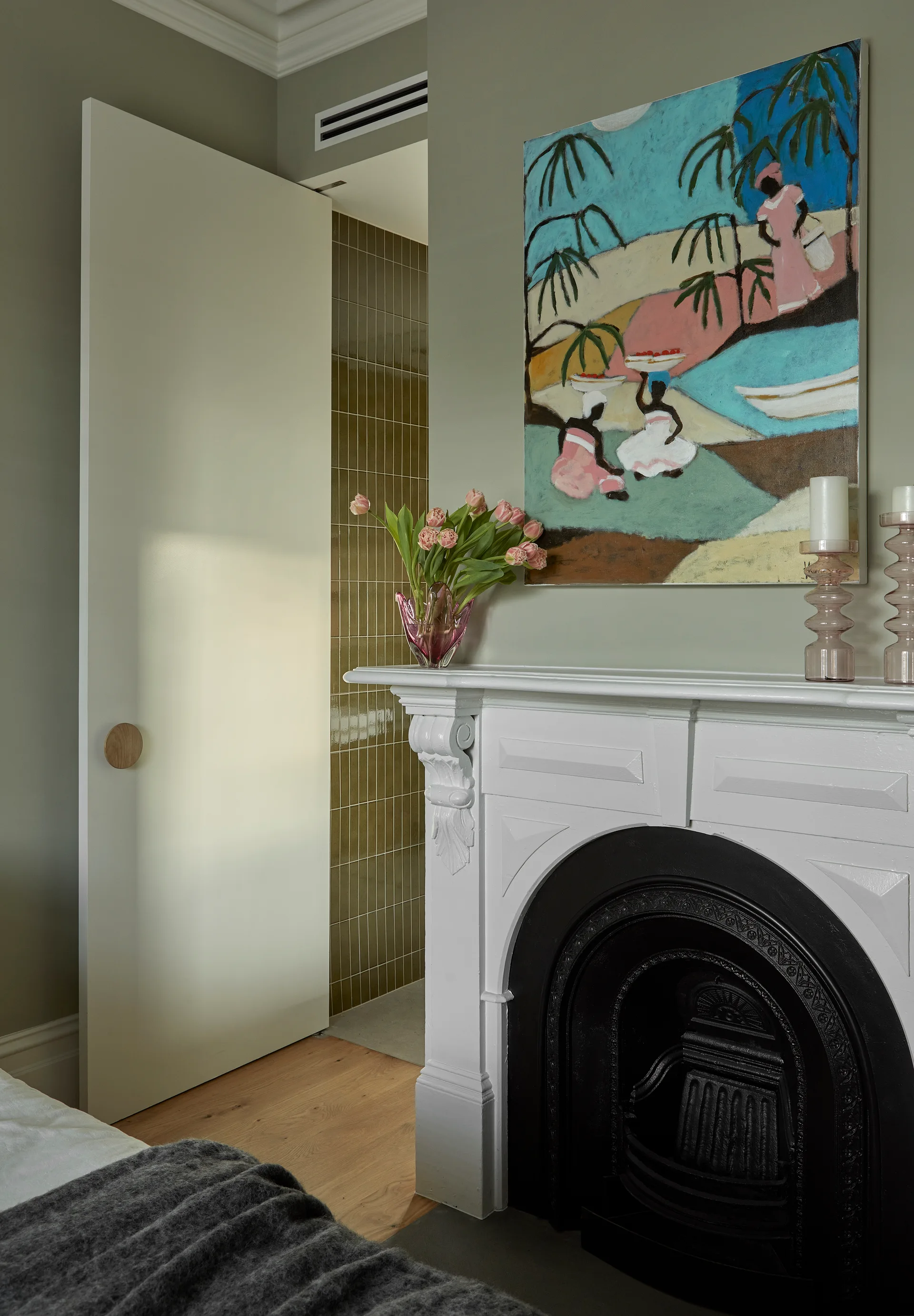 A bedroom corner features an ornate white fireplace mantel under colorful art, next to a tall white door and olive green walls.