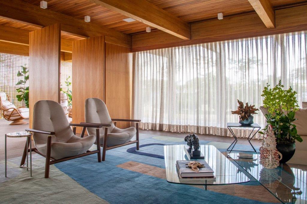 A living room featuring a wood beam ceiling, sheer curtains, two armchairs on a blue abstract rug, and a glass coffee table.