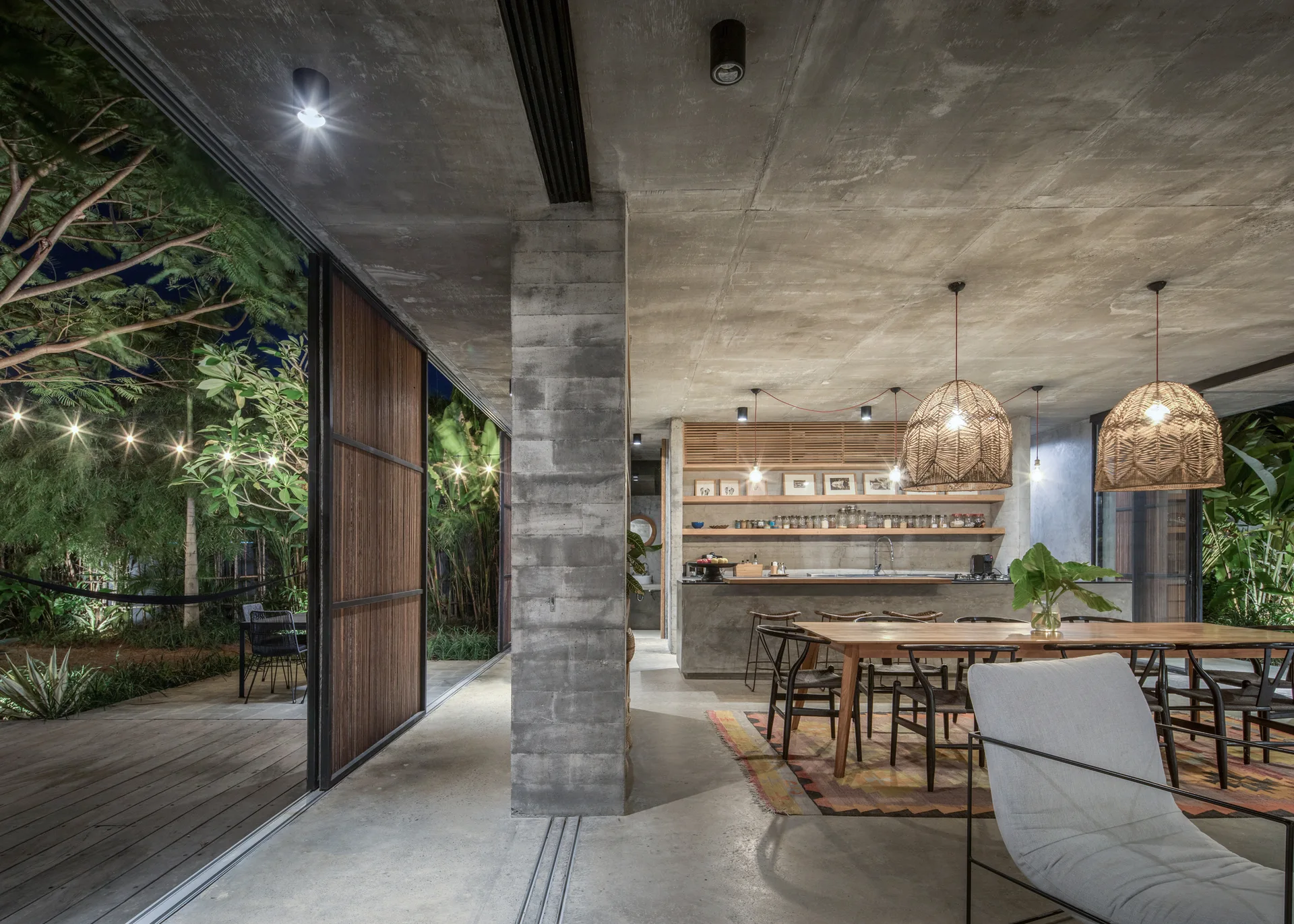 An open-concept kitchen and dining area features exposed concrete ceilings, a long wooden table with black chairs, and large woven pendant lights.
