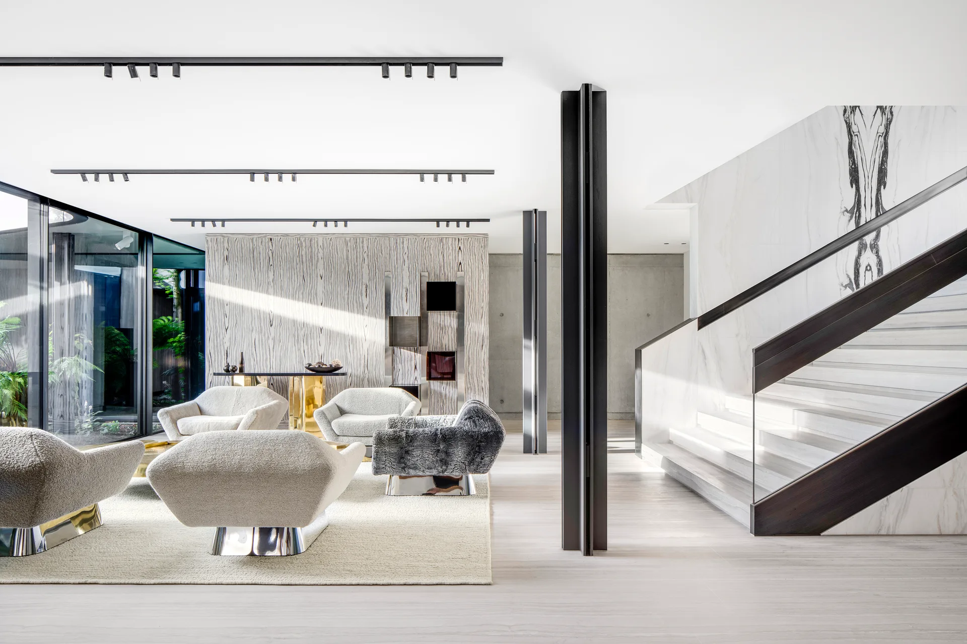 A minimalist living room features boucle armchairs with metallic bases, a marble staircase with glass railing, and black ceiling track lights.