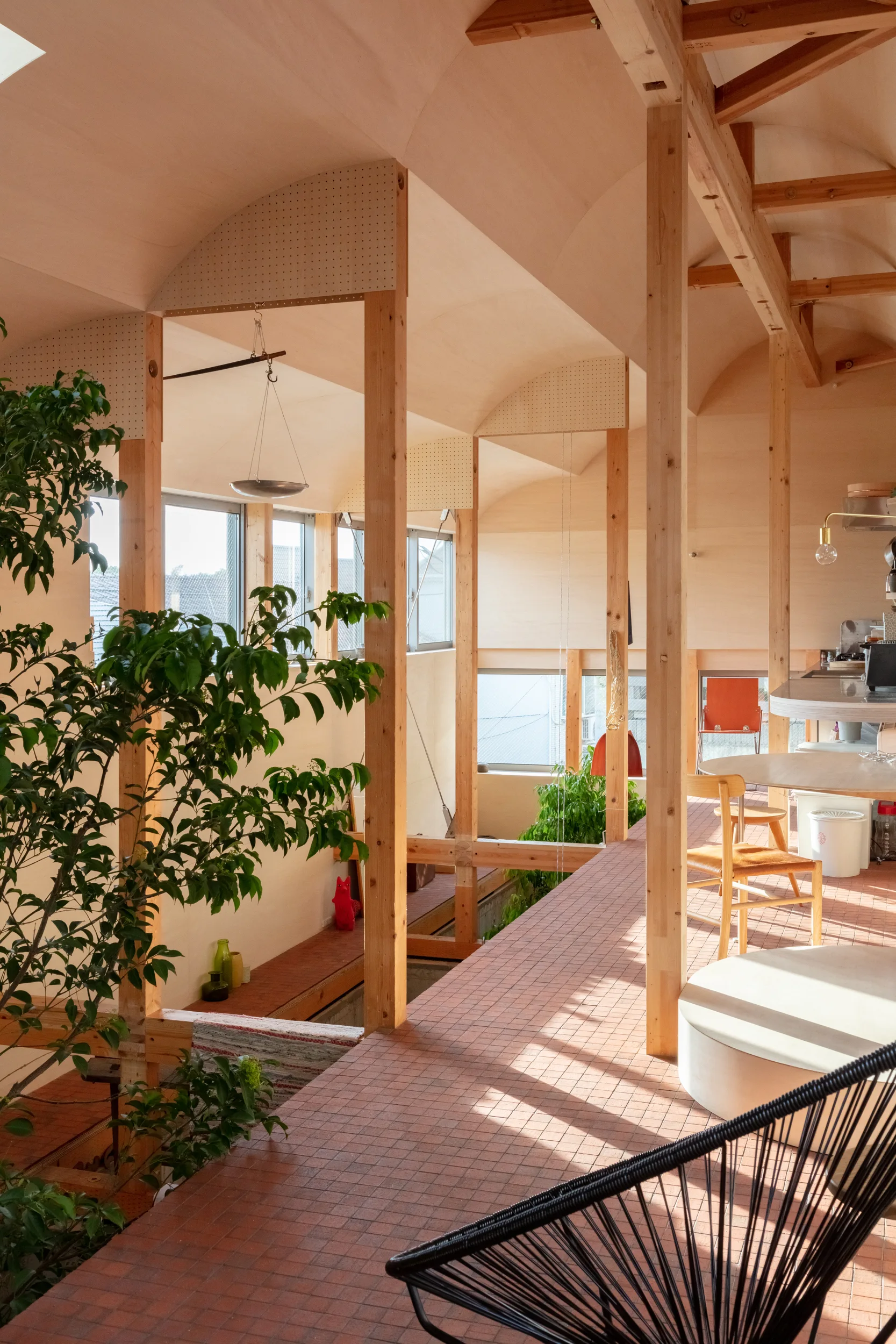 An open residential interior features an exposed timber post-and-beam frame, a vaulted plywood ceiling, and red clay grid tile flooring.