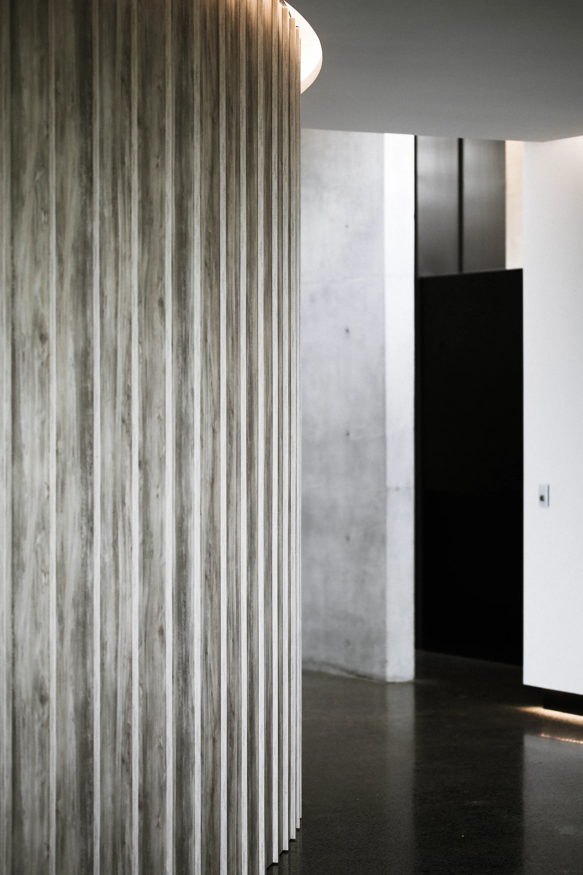 A close-up of a curved wall clad in vertical wood-toned slats under a recessed ceiling light, with a concrete wall and dark doorway behind it.