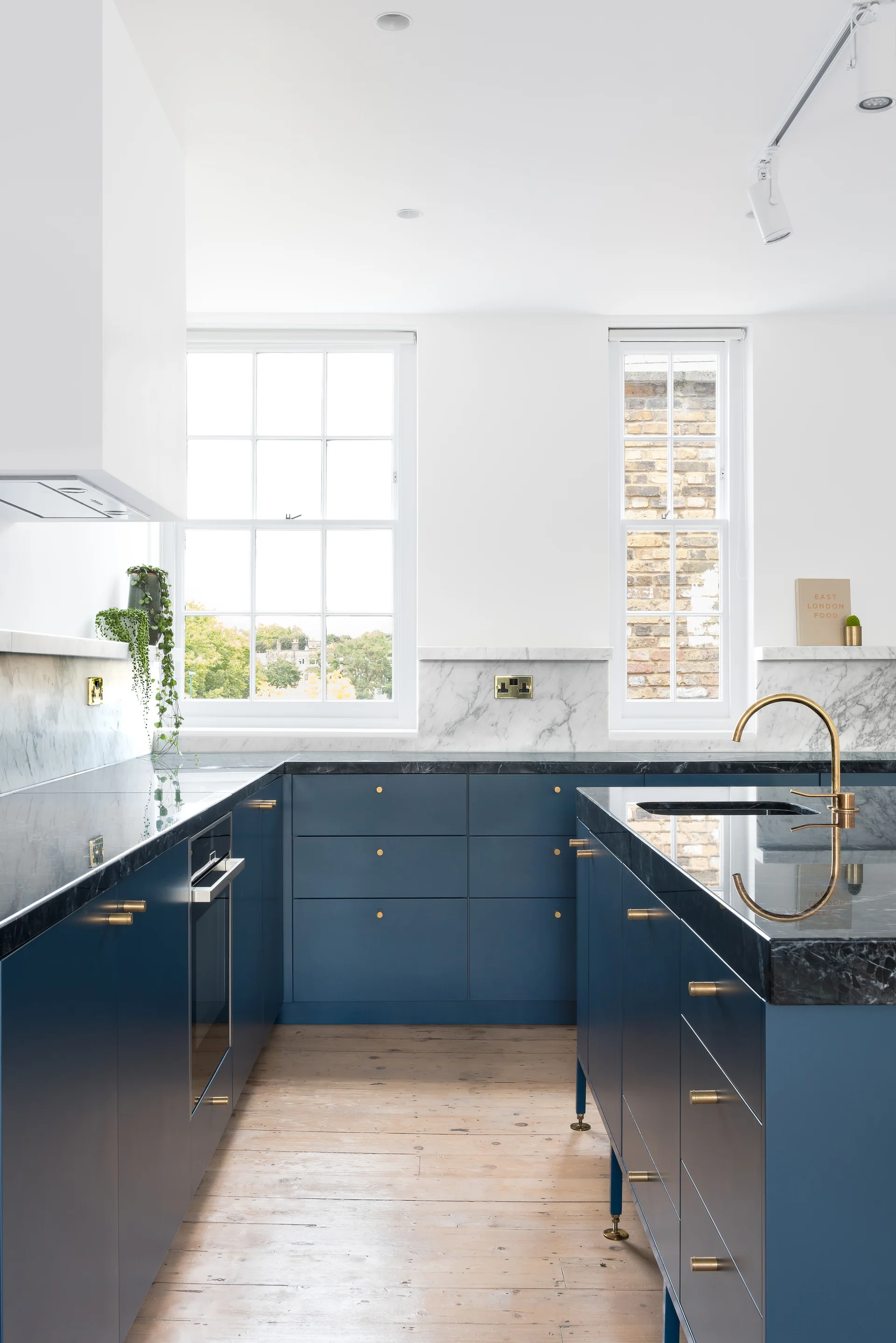 A modern kitchen features navy blue flat-panel cabinets, dark stone countertops, brass hardware, and white-framed sash windows over a wood floor.