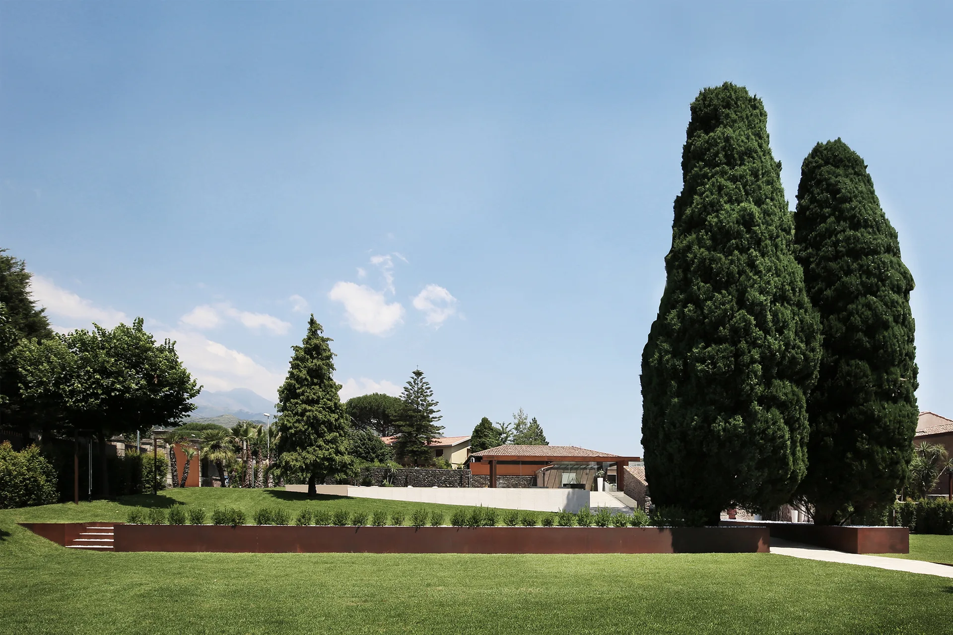 A spacious green lawn features a long rusted corten steel retaining wall planted with low shrubs, set against tall evergreen trees and a clear blue sky.