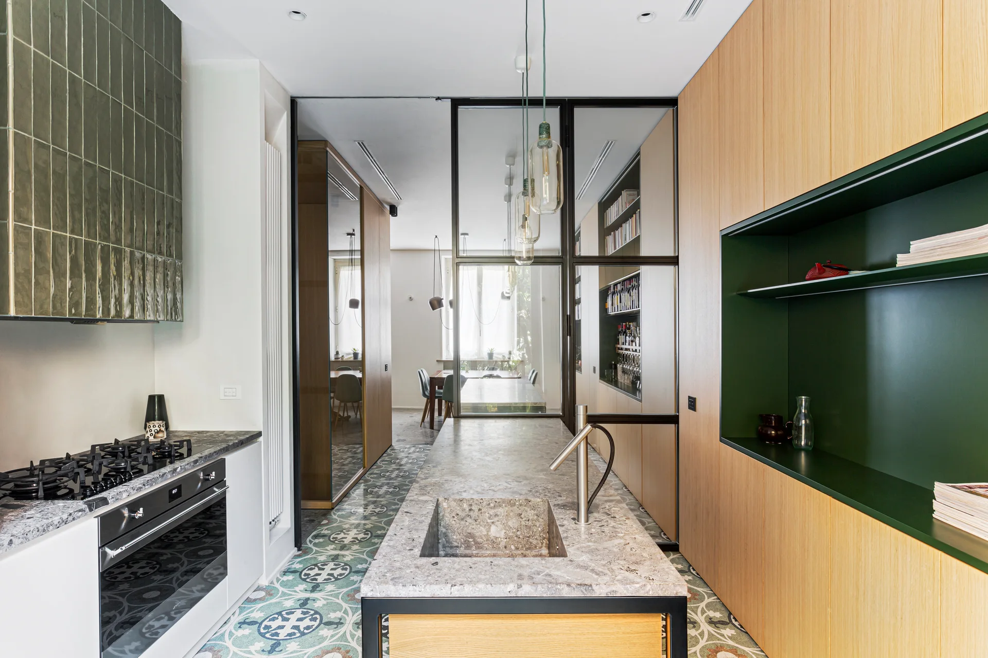 A modern kitchen featuring a stone island, light wood cabinetry, a dark green recessed shelf, patterned floor tiles, and a black-framed glass partition.