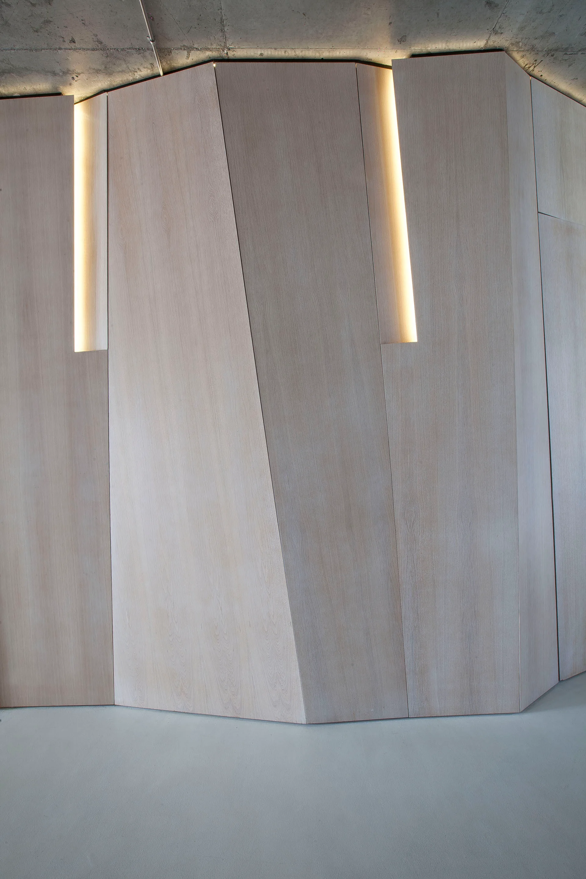 Asymmetrical light wood wall panels feature integrated vertical glowing light strips beneath a raw exposed concrete ceiling edge.