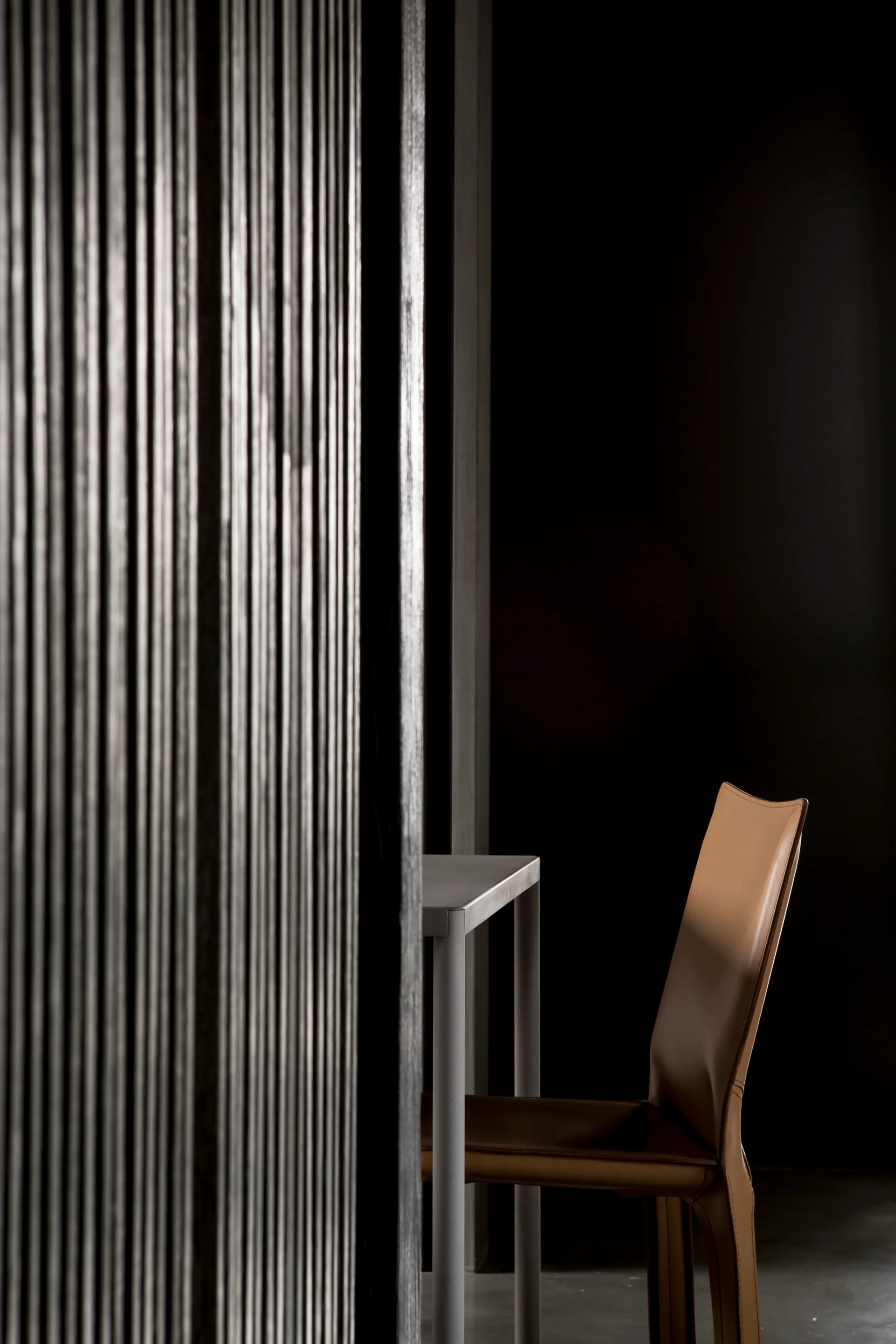 A minimalist brown leather chair and grey metal table stand next to a dark vertical slatted wall partition.