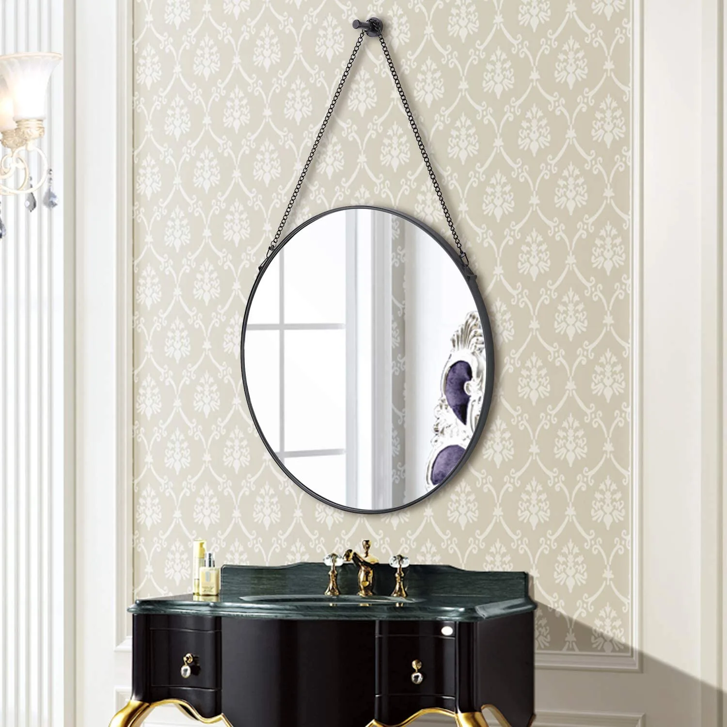 A round black-framed mirror hangs from a delicate chain on cream damask wallpaper above a dark marble-topped vanity.