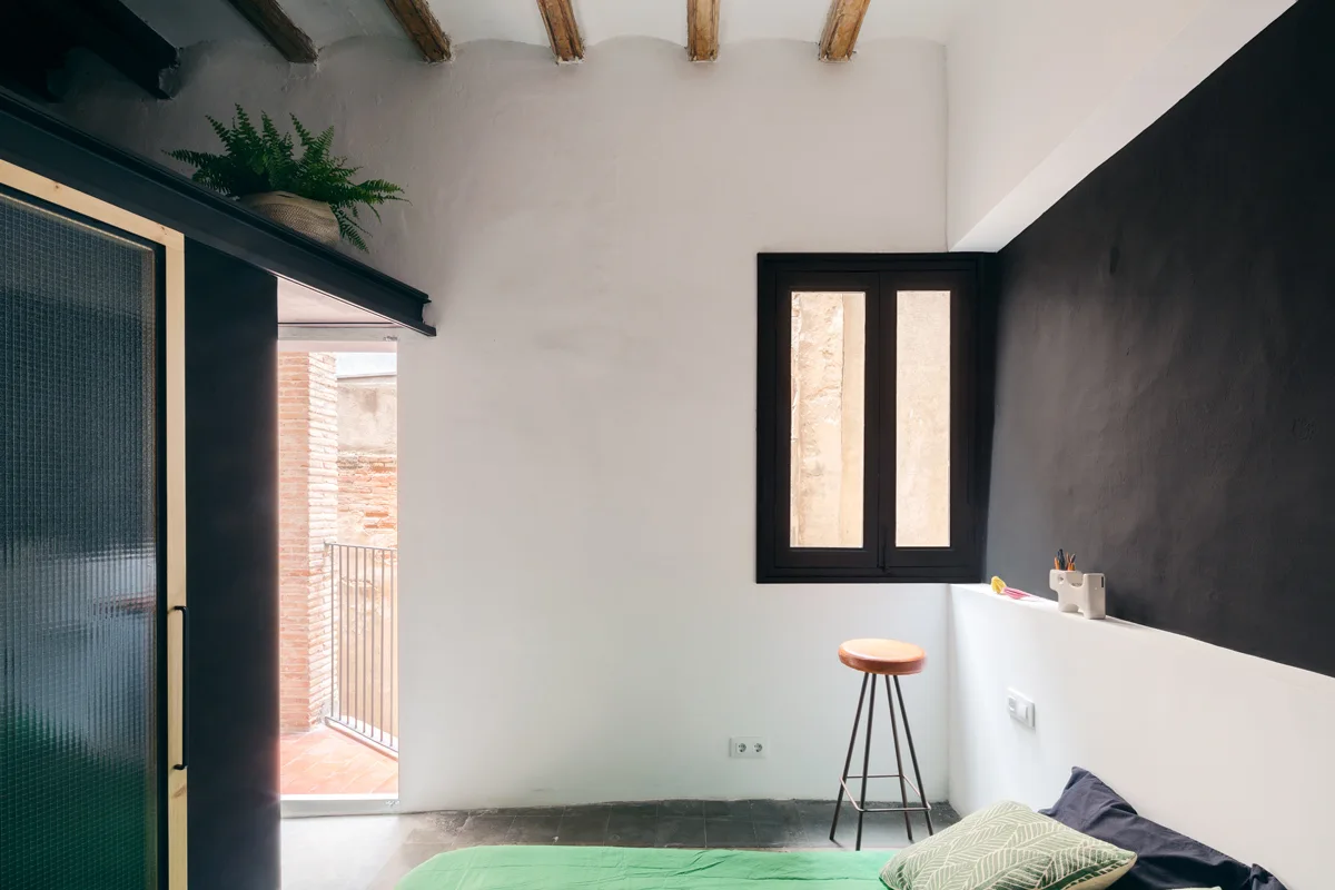 A minimalist bedroom features a bright green bed, a black accent wall, exposed wooden ceiling beams, and a small black-framed window.