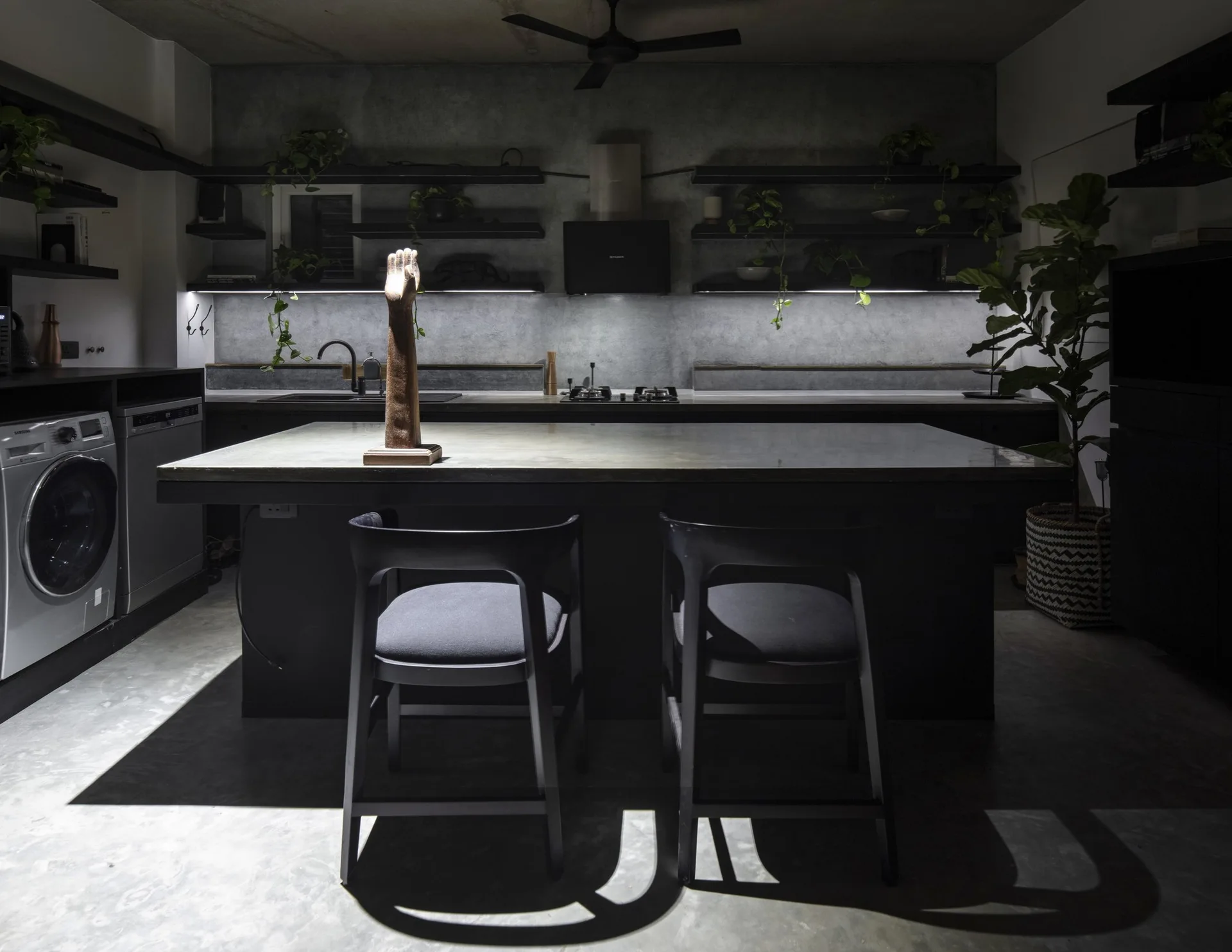 A dark industrial kitchen featuring a large black island with two chairs, textured concrete walls, and open shelving with integrated strip lighting.