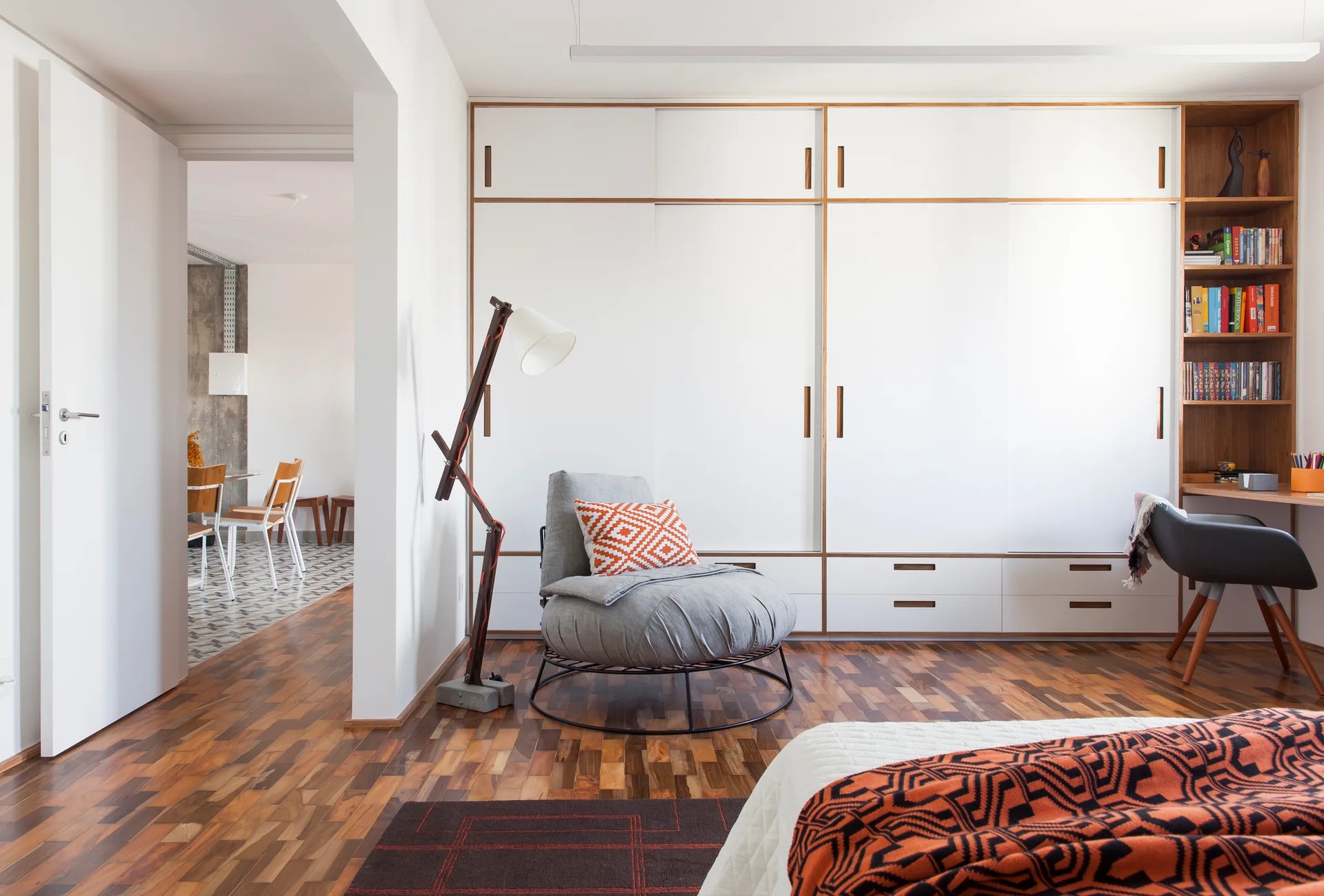 A modern bedroom features a large white built-in wardrobe with wood trim, parquet flooring, a gray circular armchair, and an integrated desk.
