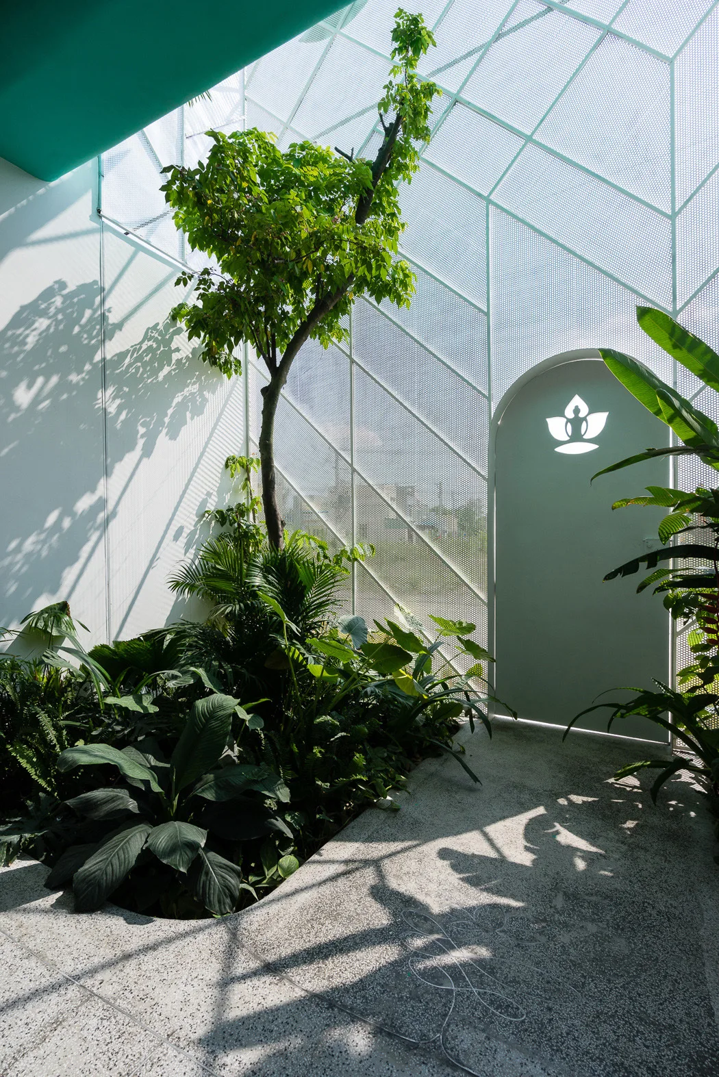 An indoor garden atrium features a central tree, lush green plants, speckled concrete flooring, and a perforated white metal mesh wall with an arched door.