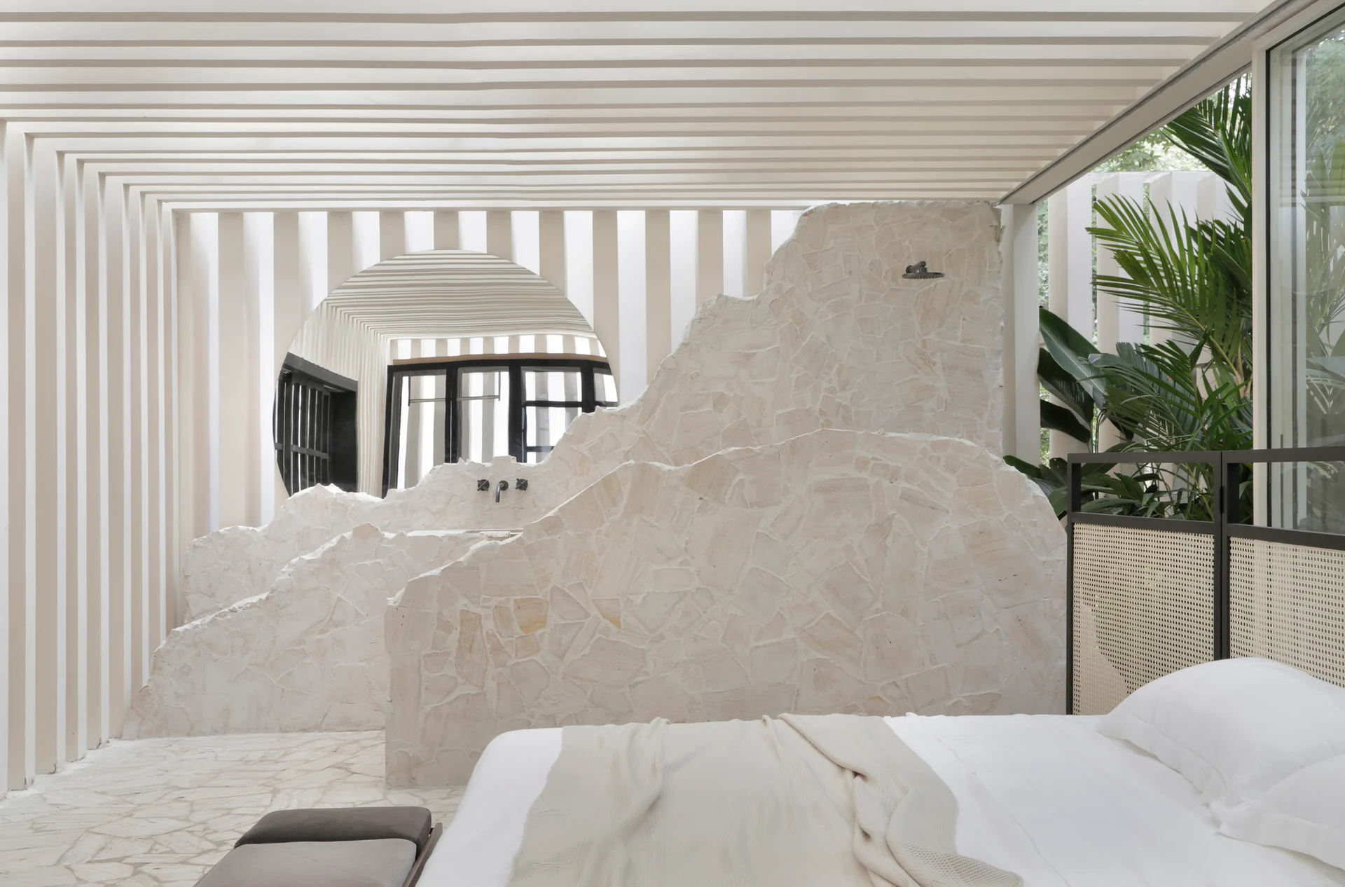 A modern bedroom featuring a jagged stone shower partition wall next to a bed with a white comforter and rattan headboard.