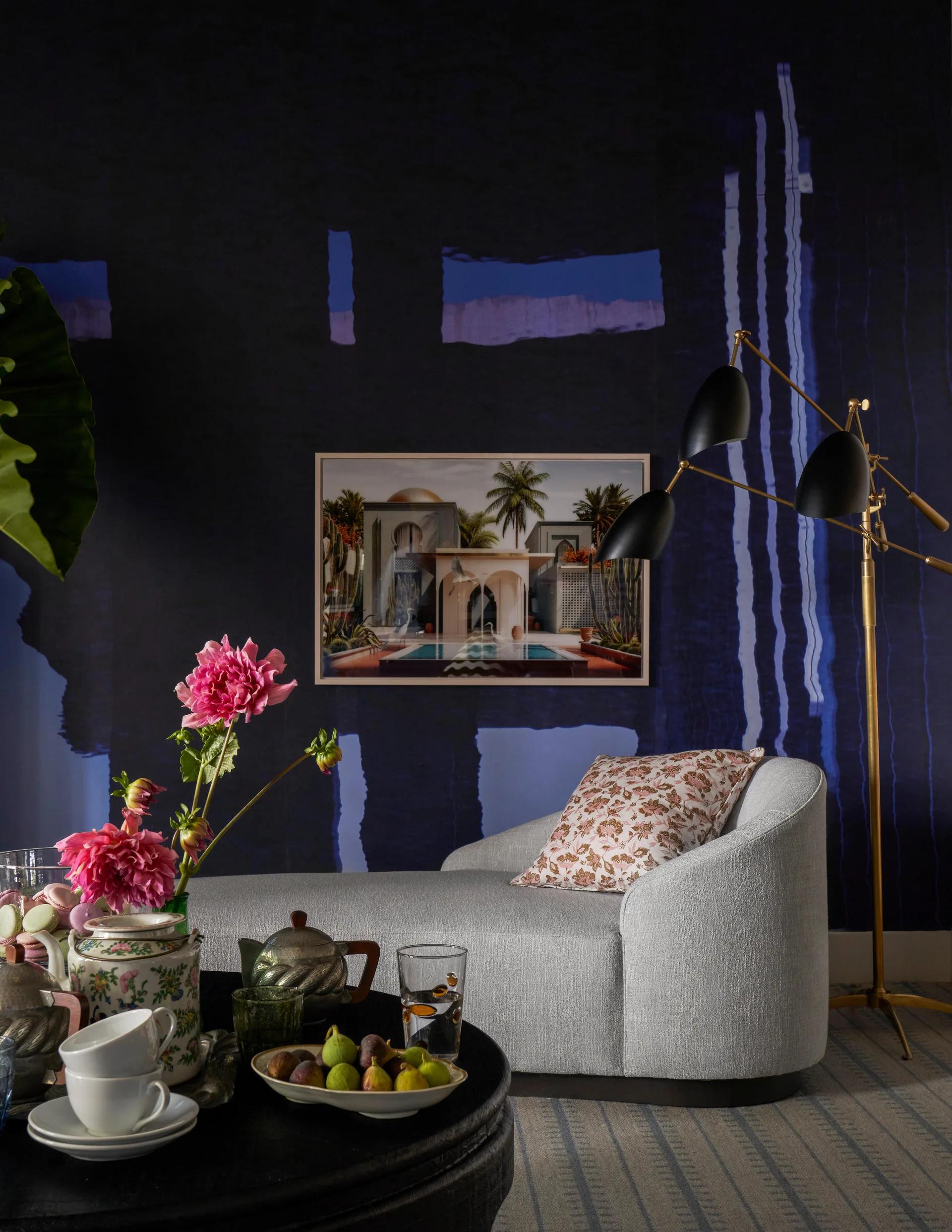 A gray upholstered chaise lounge sits against a dark blue patterned wall next to a three-arm gold floor lamp and a coffee table with tea service.