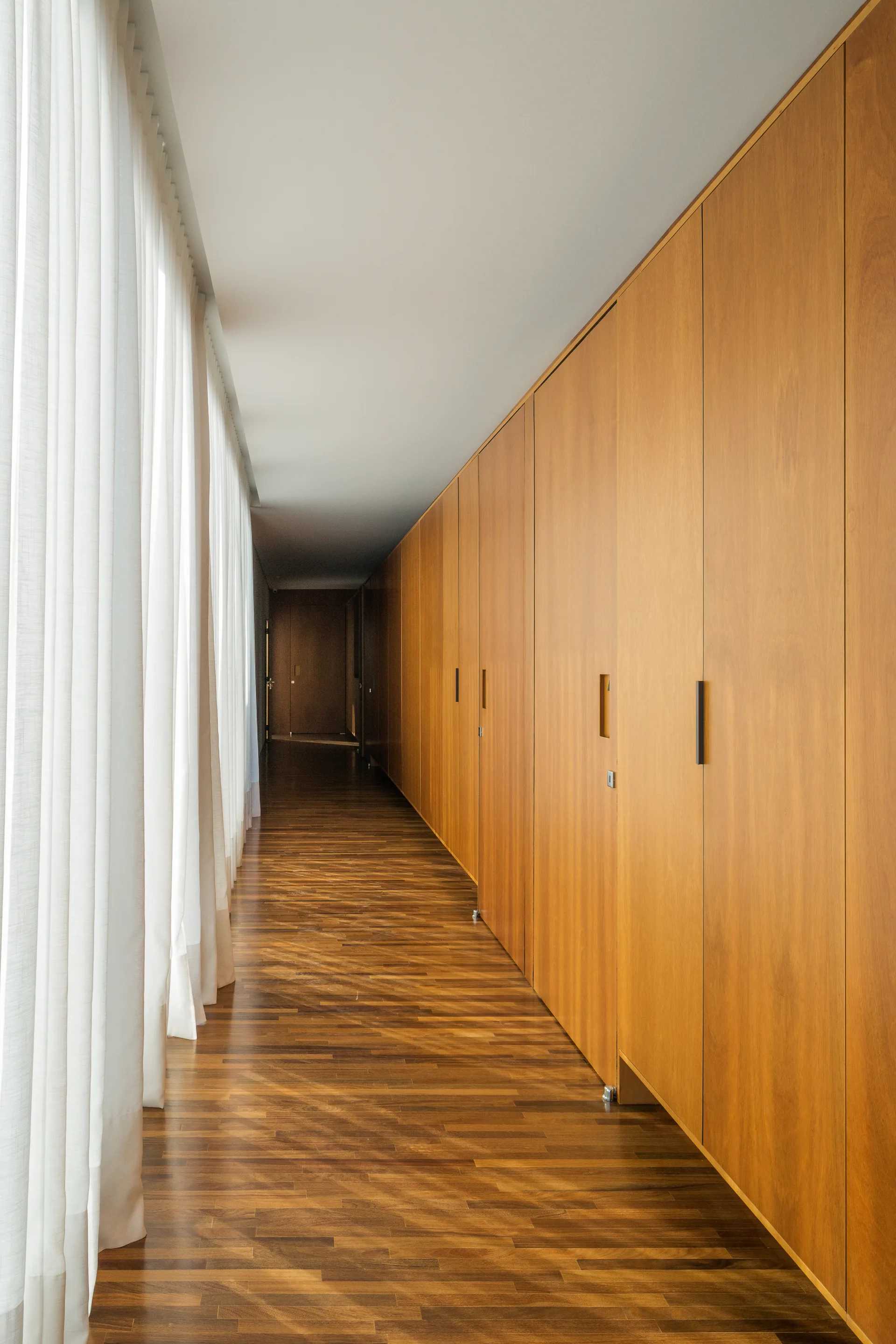 A long hallway with warm wood flooring features built-in flat-panel wood closets on one side and sheer white curtains on the other.