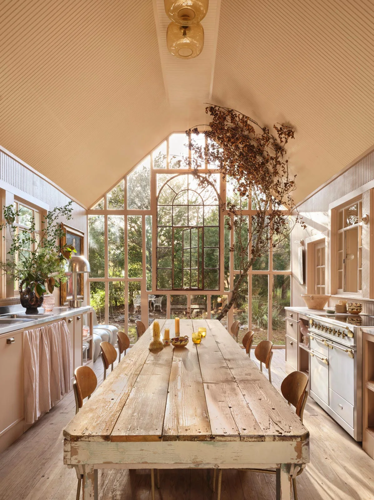 A rustic kitchen and dining area features a long weathered wood table, a vaulted beadboard ceiling, and a large arched window looking out at trees.