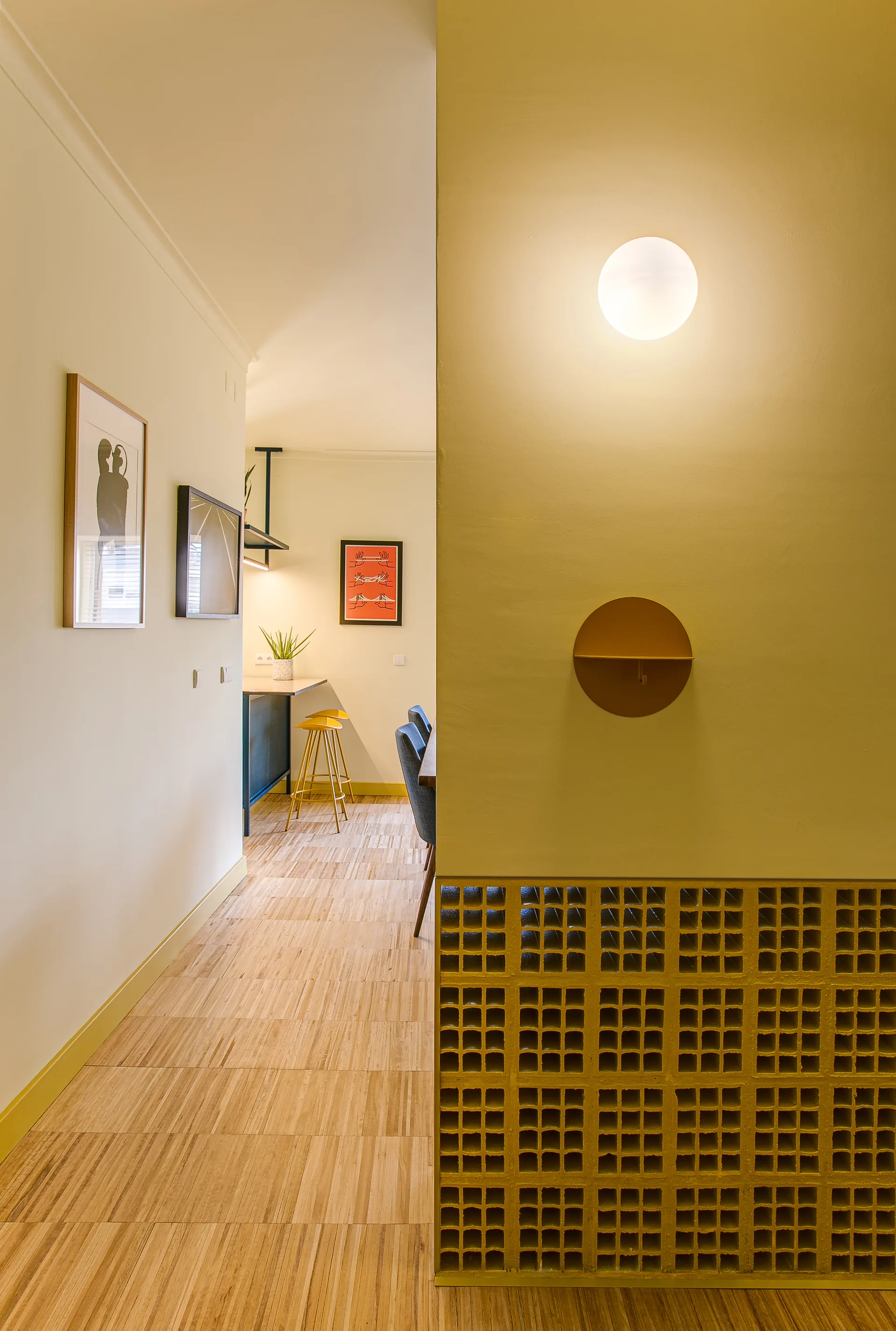 A hallway with light wood flooring and a cream wall leading to a kitchen, featuring a partition wall detailed with yellow breeze blocks and a round sconce.