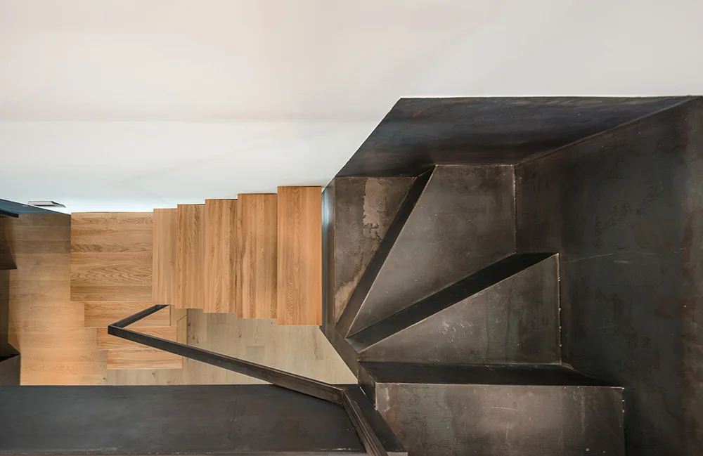 An overhead view of a modern staircase featuring light wood treads juxtaposed with a dark metal landing and a thin black handrail.