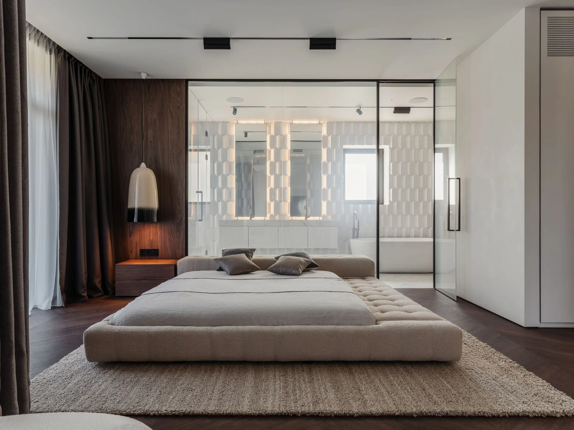 A contemporary bedroom features a low tufted platform bed resting on a textured rug in front of a clear glass wall revealing an ensuite bathroom.