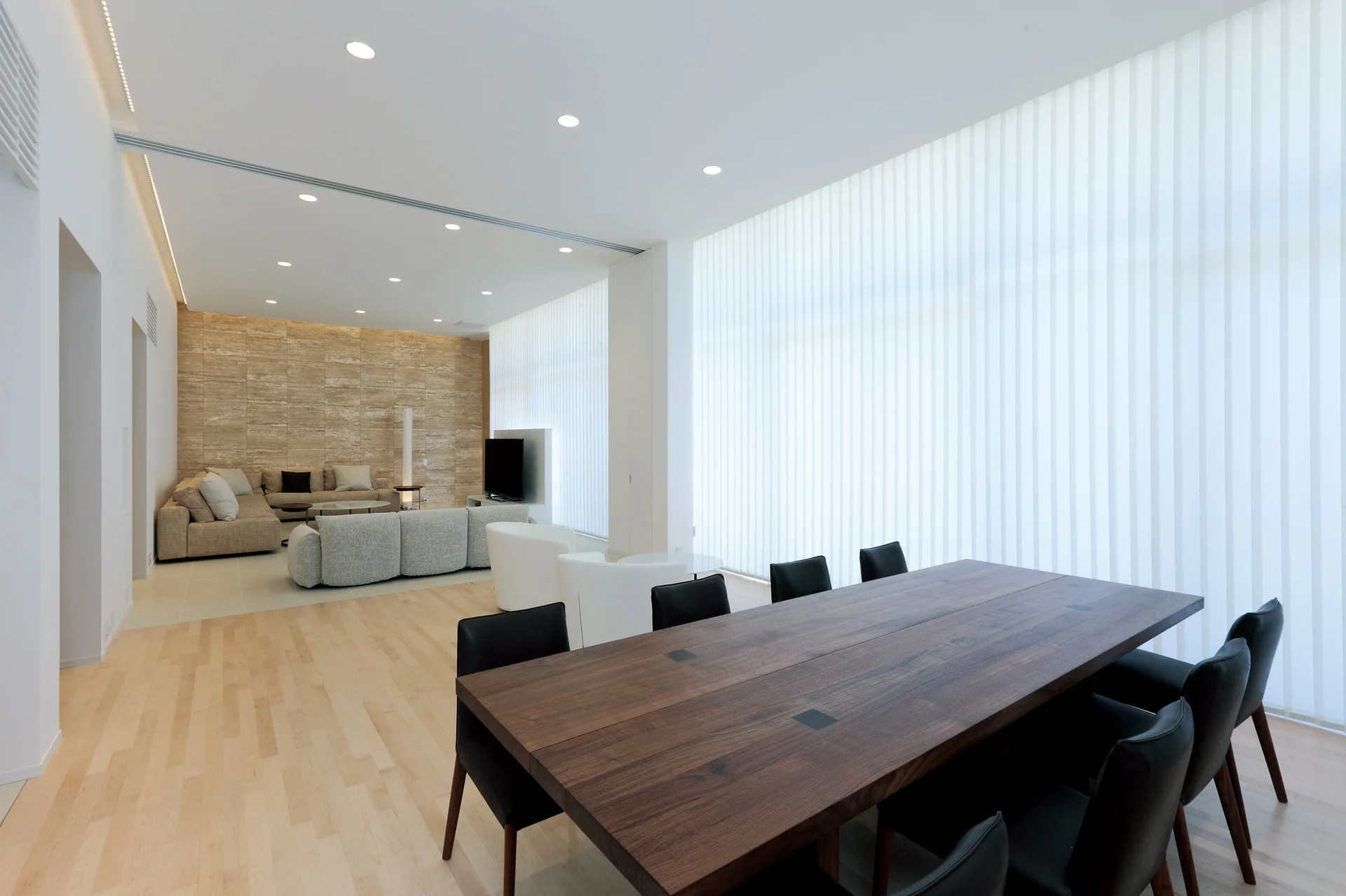 An open-concept room features a large dark wood dining table with black chairs, leading into a living space with a textured beige stone accent wall.