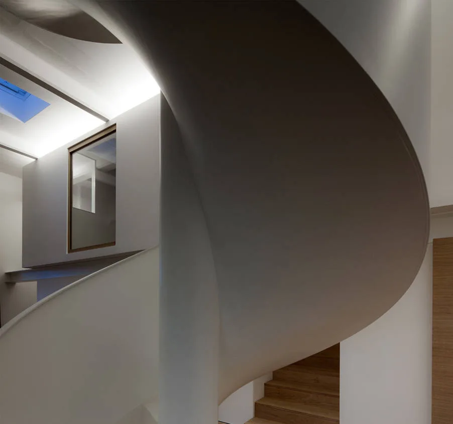 A white minimalist spiral staircase twists upward toward a skylight, showing natural wood steps and warm cove lighting on the walls.
